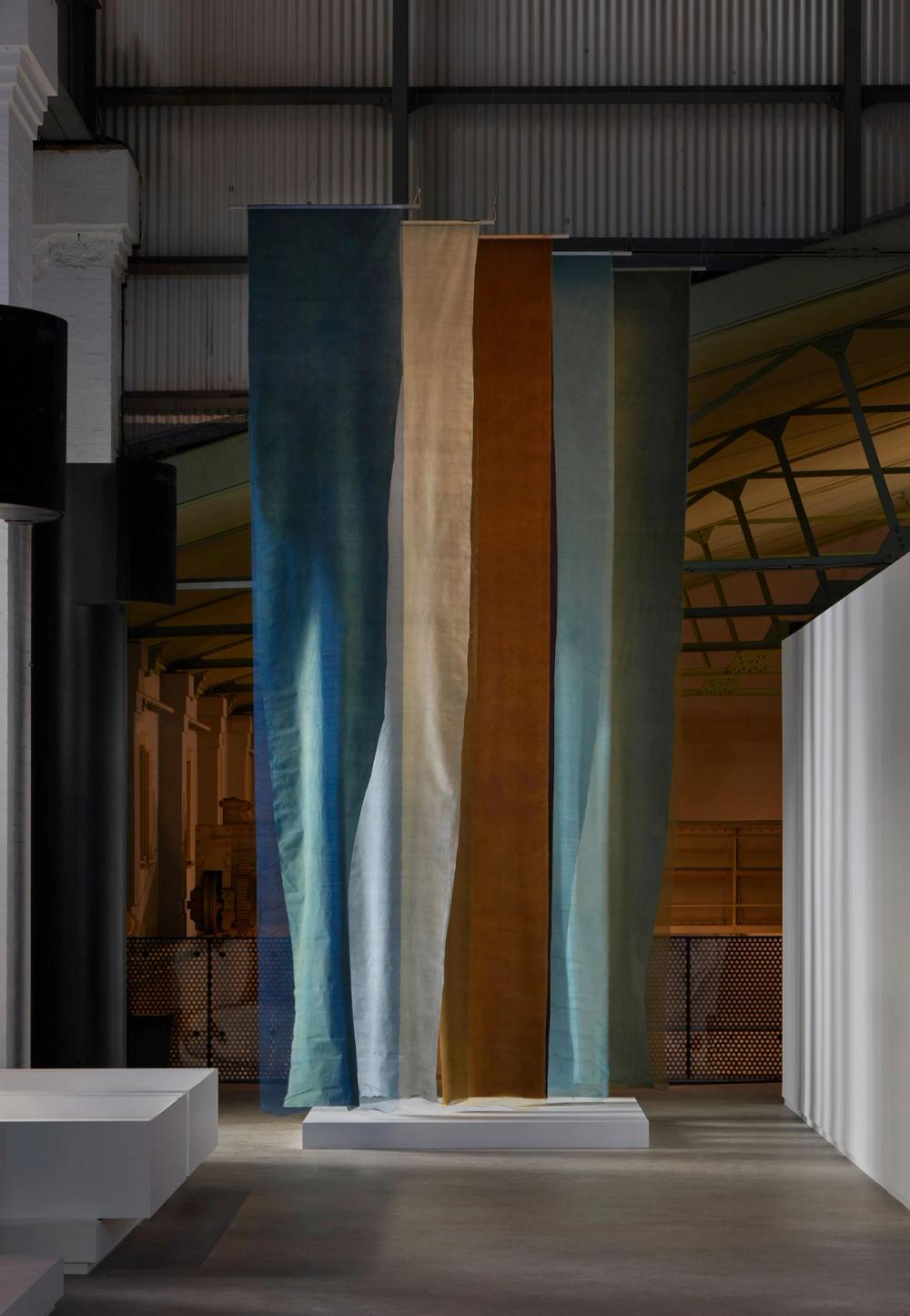 Multiple lengths of textile hang above a low white plinth, the textiles are shades of blue, beige and orange.