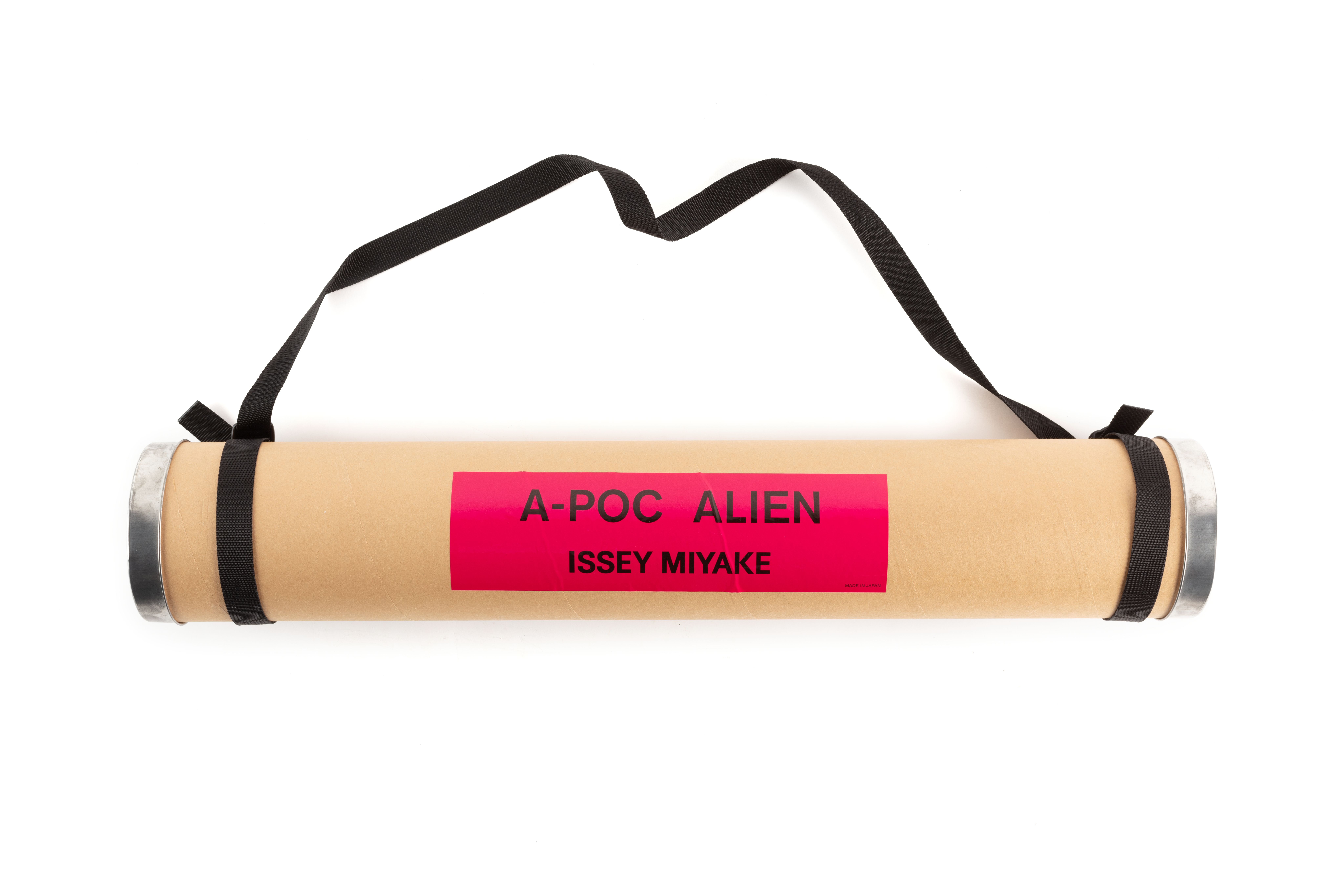Brown cardboard tube with pink and black sticker 'A-POC ALIEN/ISSEY MIYAKE/MADE IN JAPAN' and two silver metal lids. Looped around each end of the tube is a black nylon webbing carry strap fastened with black plastic buckles.