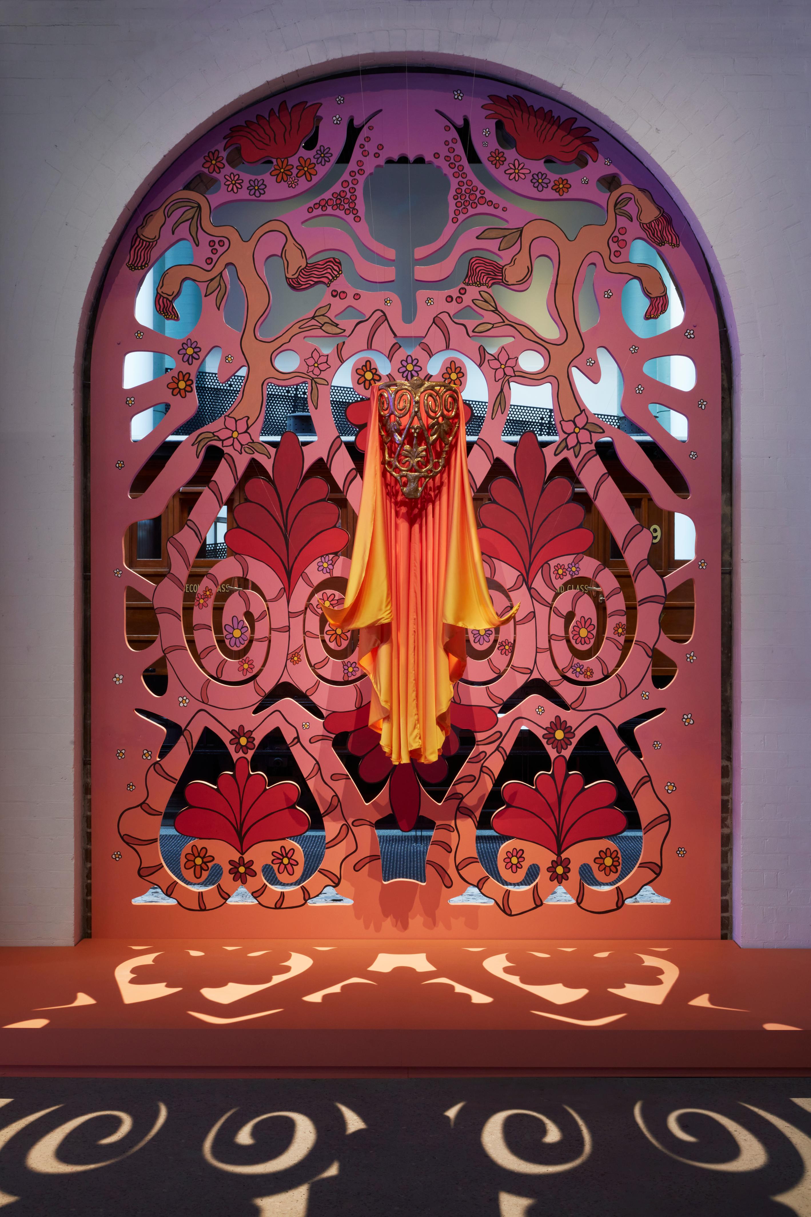 A large peach, pink, and orange archway panel with cut out panel. In front a cap and body piece hangs as if levitating.