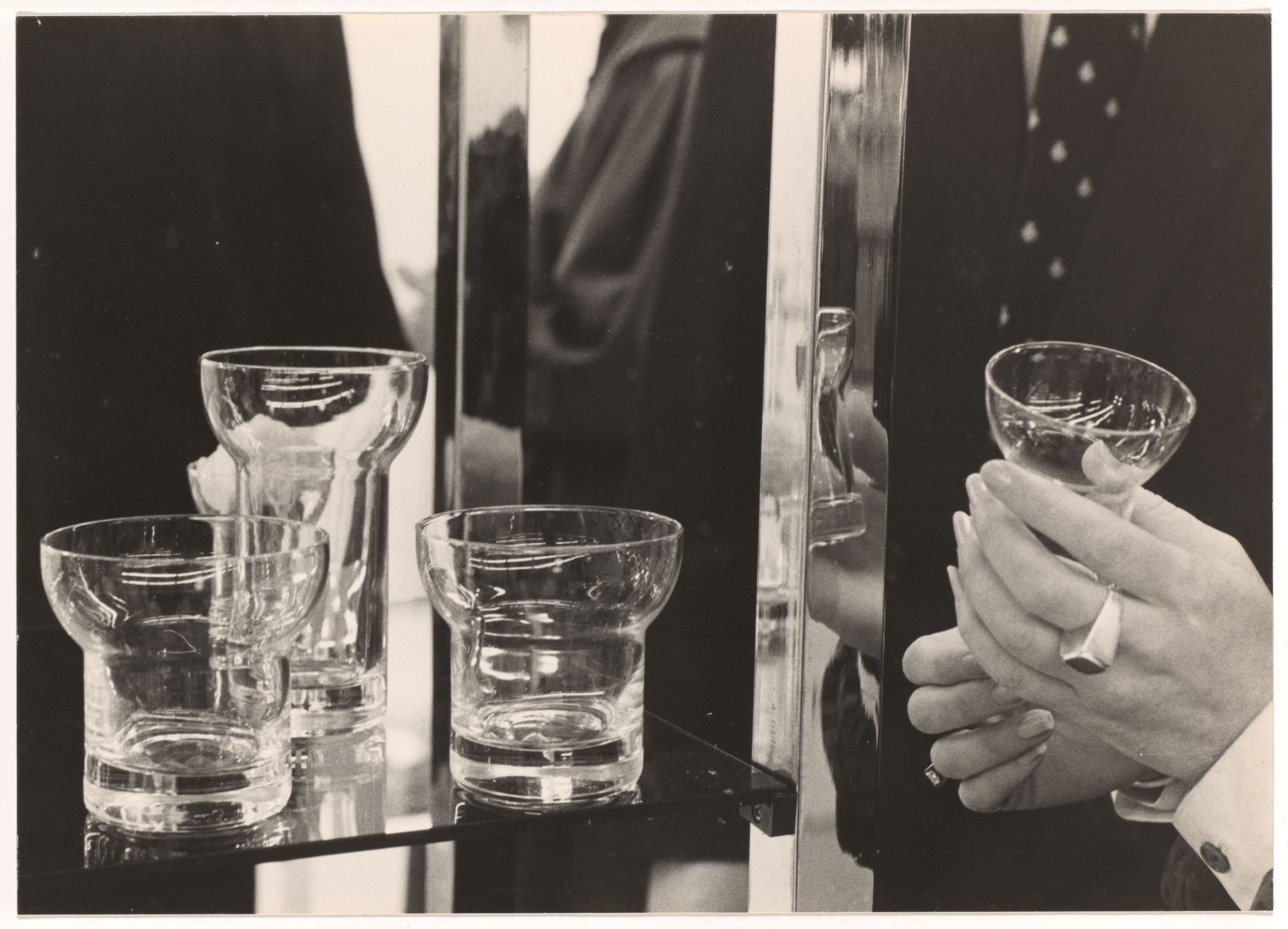 Black and white close up photograph of four curved glasses on a shelf, one being held by designer Denise Larcombe.