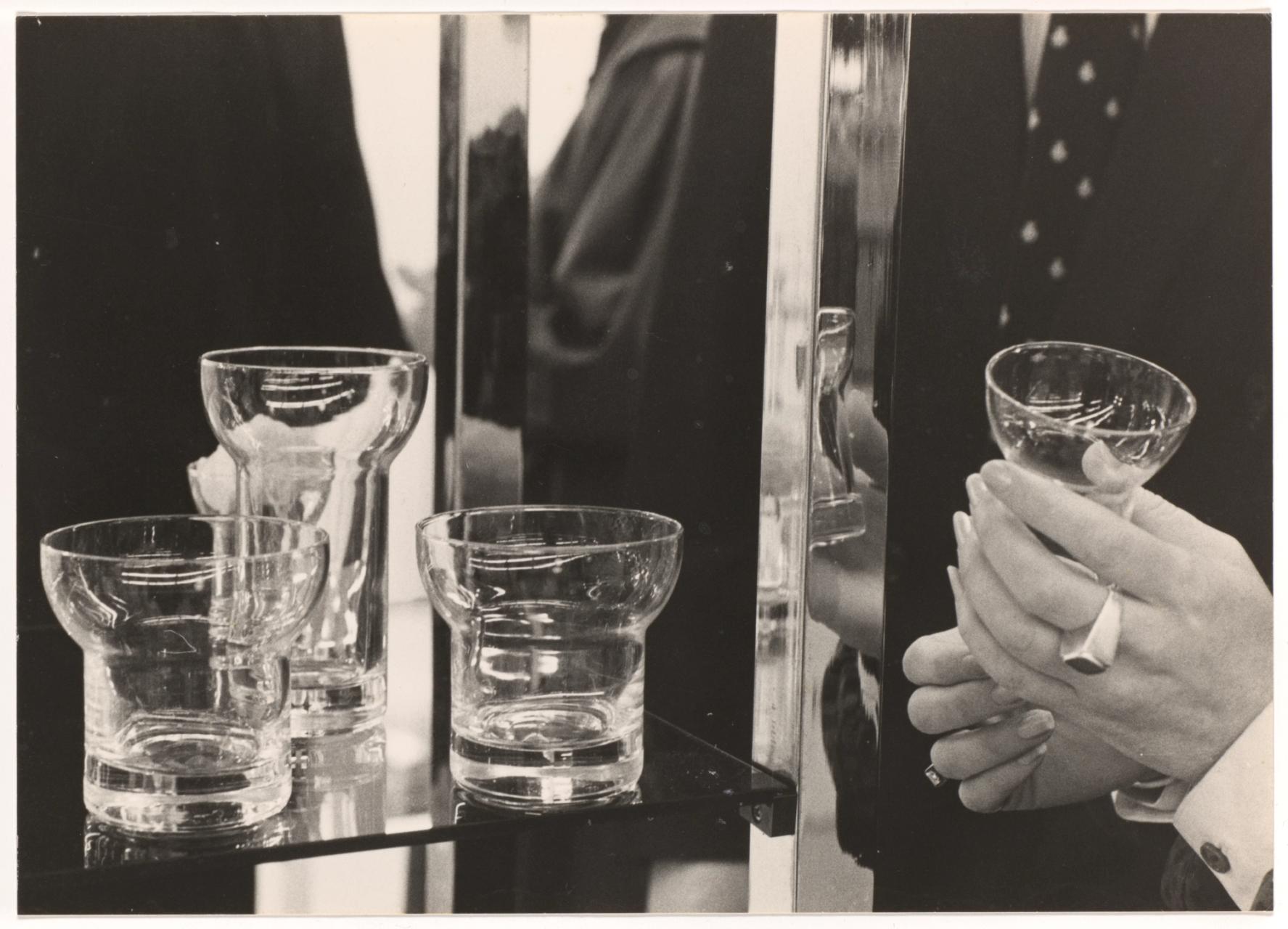 Black and white close up photograph of four curved glasses on a shelf, one being held by designer Denise Larcombe.