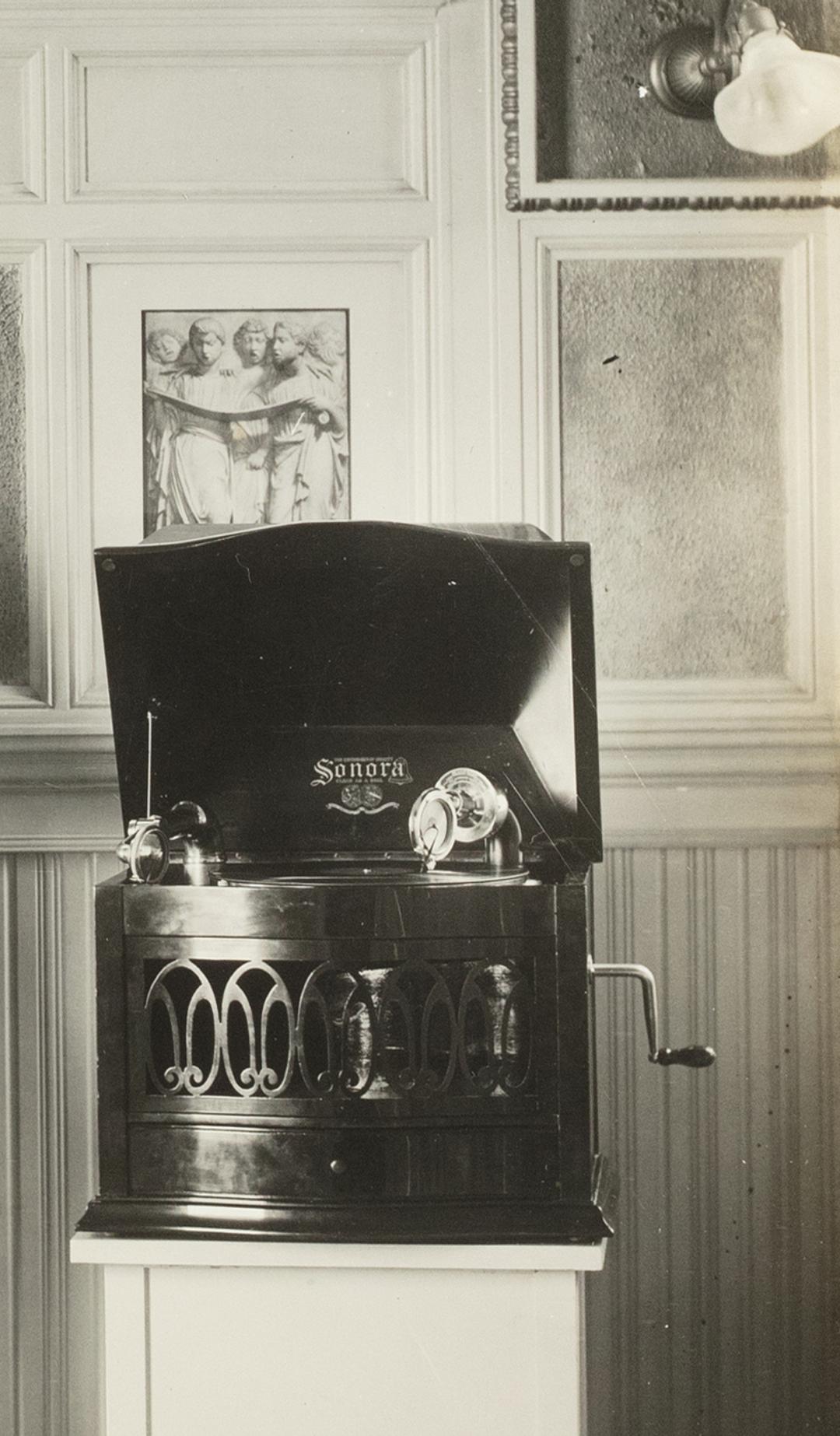 A black and white photographic print mounted on a sheet of linen. Photo of Captain Sharp demonstrating two Magnavox phonographs, white paper label glued to back 'CAPTAIN SHARP DEMONSTRATING WITH MV-1 & SONORA'.