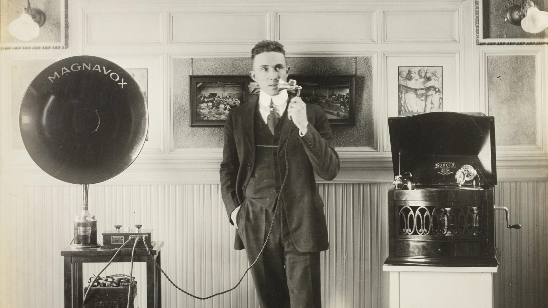 A black and white photographic print mounted on a sheet of linen. Photo of Captain Sharp demonstrating two Magnavox phonographs, white paper label glued to back 'CAPTAIN SHARP DEMONSTRATING WITH MV-1 & SONORA'.