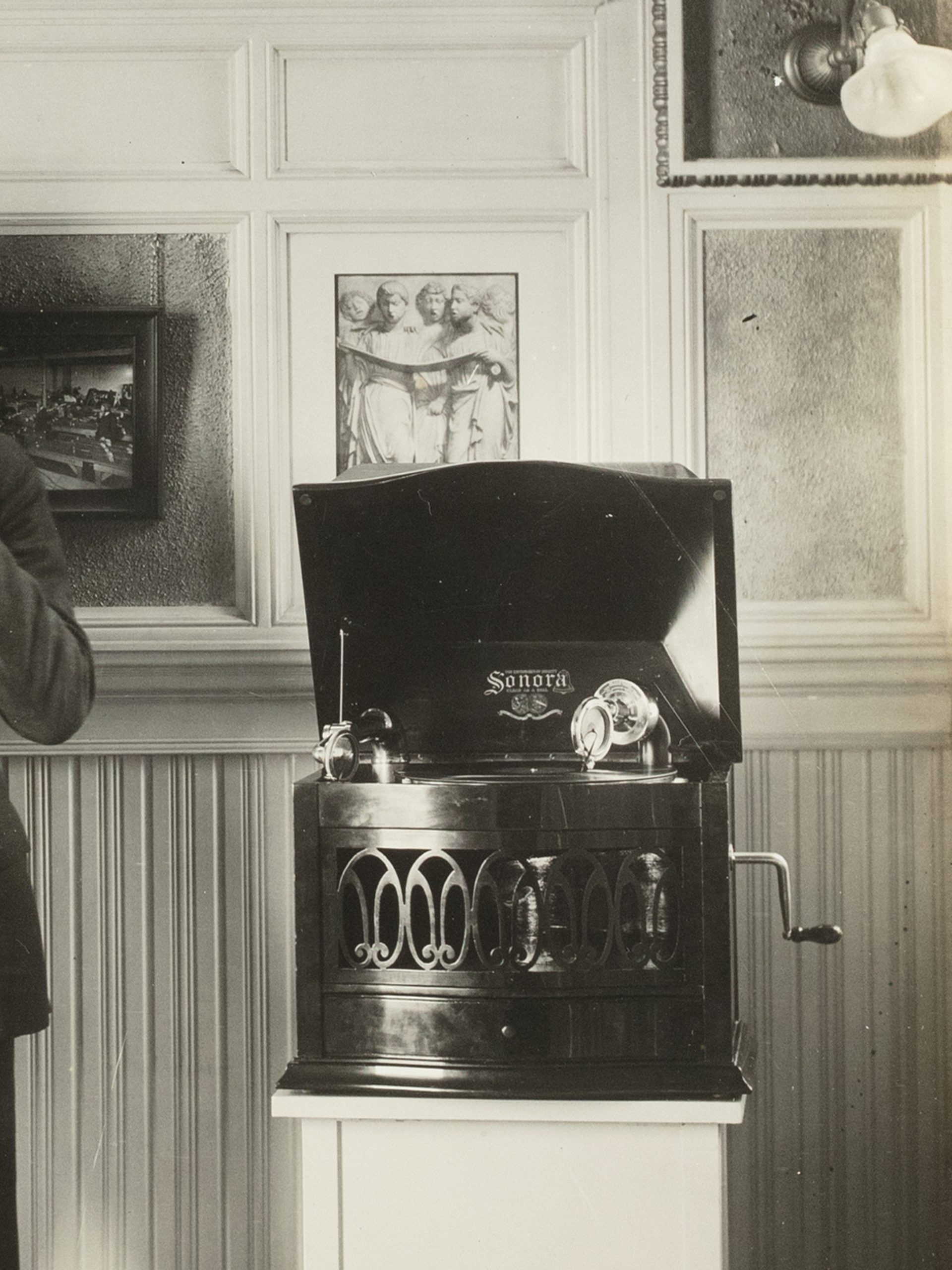 A black and white photographic print mounted on a sheet of linen. Photo of Captain Sharp demonstrating two Magnavox phonographs, white paper label glued to back 'CAPTAIN SHARP DEMONSTRATING WITH MV-1 & SONORA'.