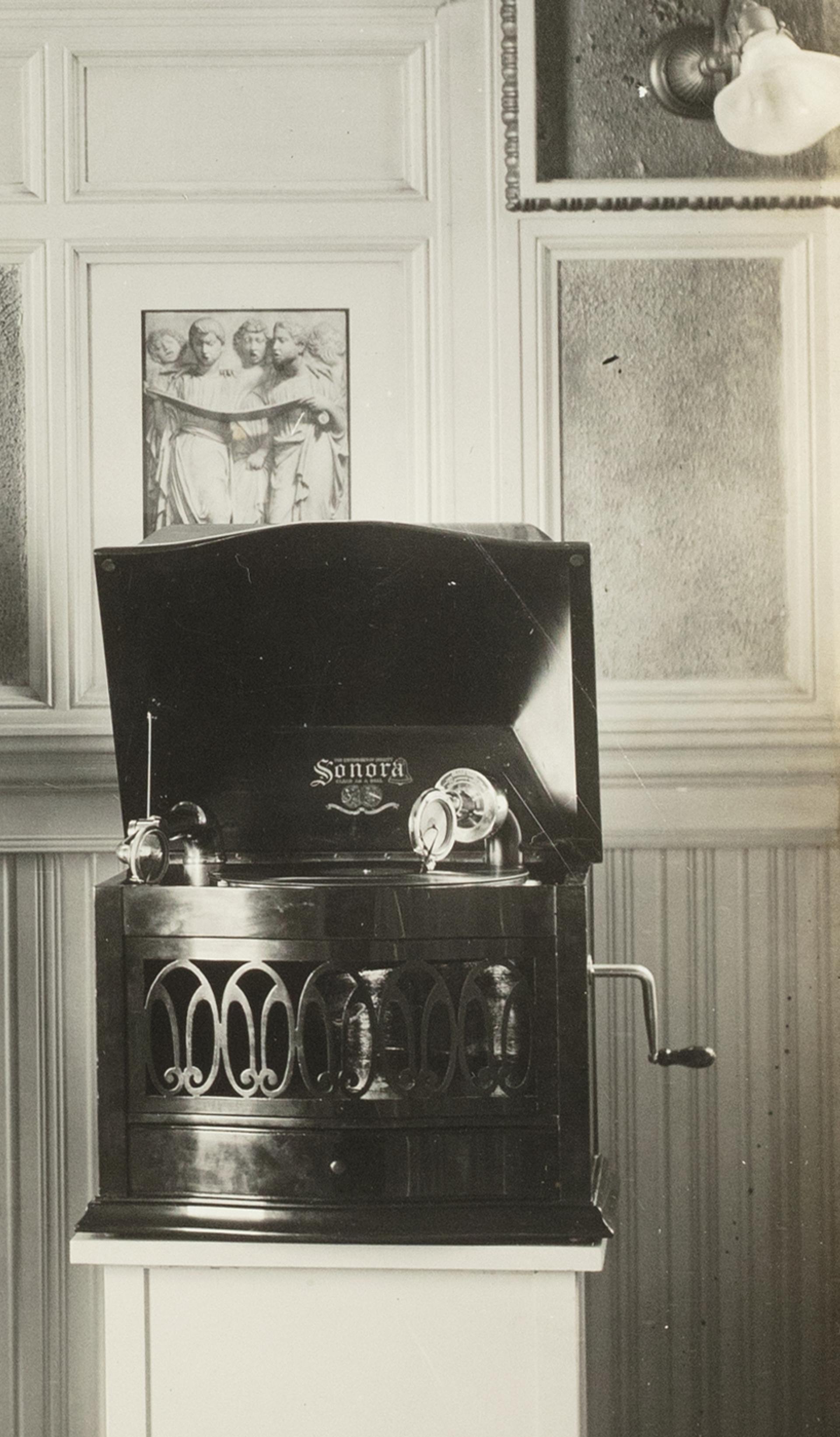 A black and white photographic print mounted on a sheet of linen. Photo of Captain Sharp demonstrating two Magnavox phonographs, white paper label glued to back 'CAPTAIN SHARP DEMONSTRATING WITH MV-1 & SONORA'.