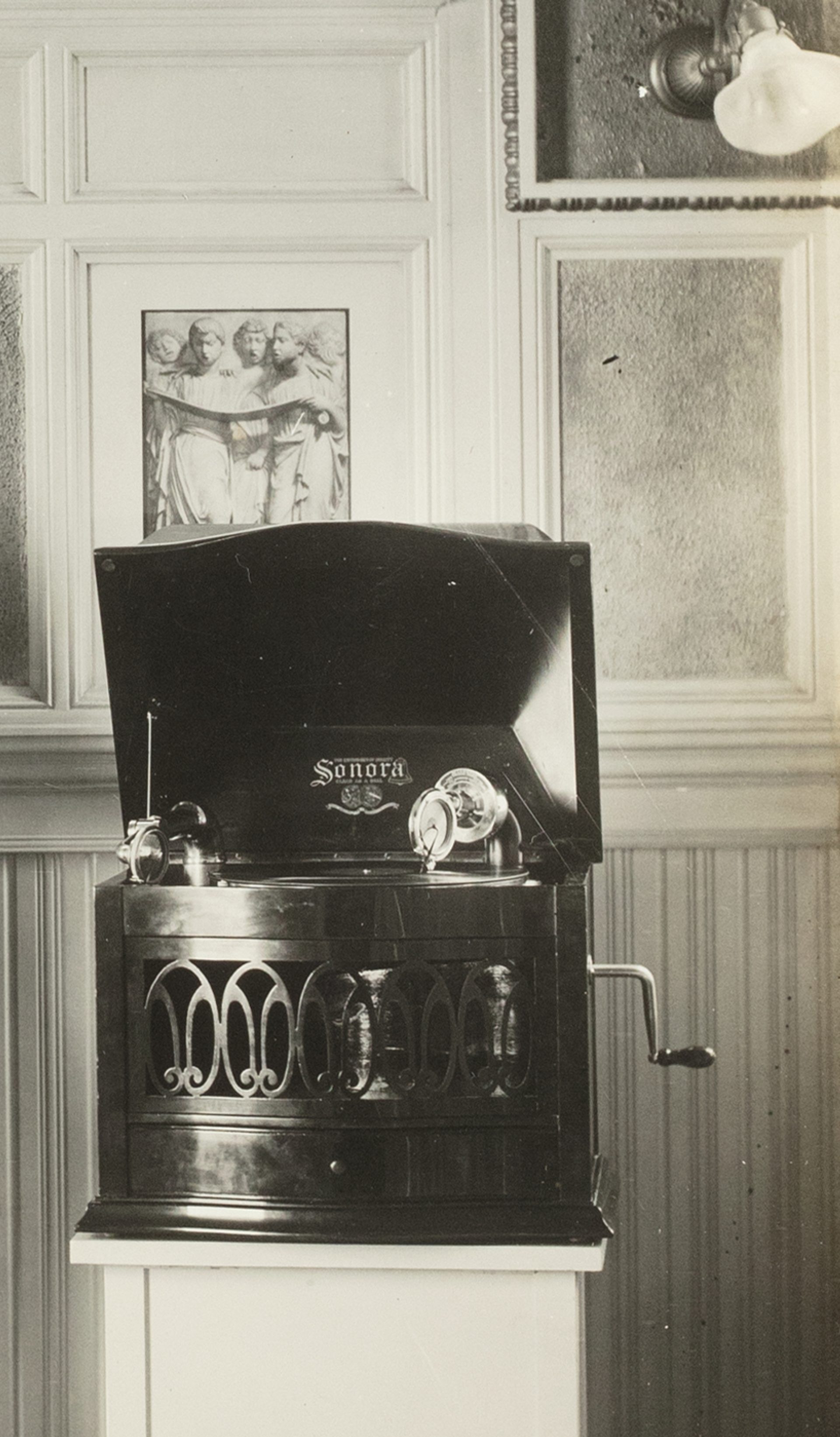 A black and white photographic print mounted on a sheet of linen. Photo of Captain Sharp demonstrating two Magnavox phonographs, white paper label glued to back 'CAPTAIN SHARP DEMONSTRATING WITH MV-1 & SONORA'.