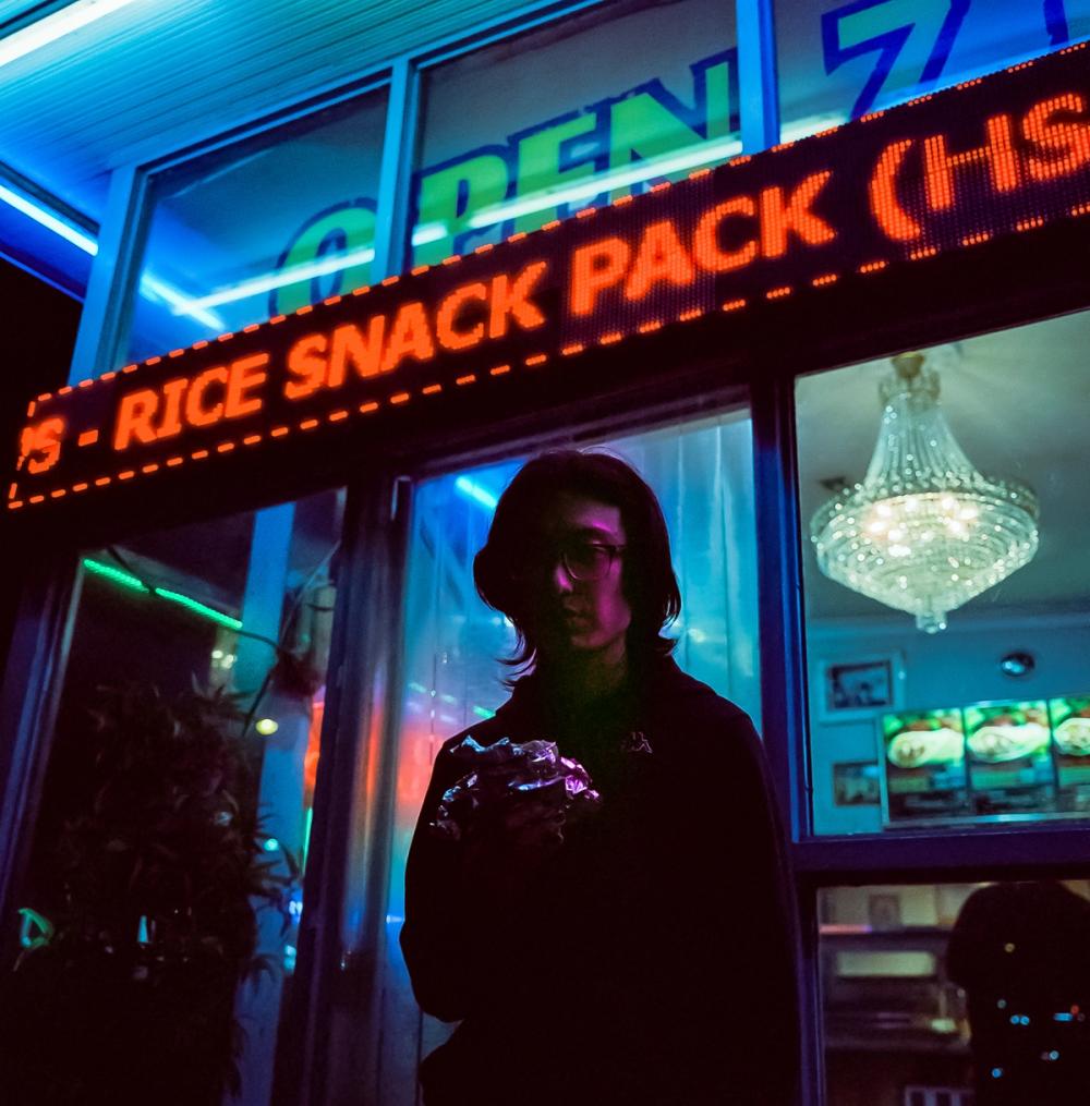 A young man stands outside a kebab store at night. He has shoulder length black hair, wears a black hoodie and is holding a kebab wrapped in silver foil.