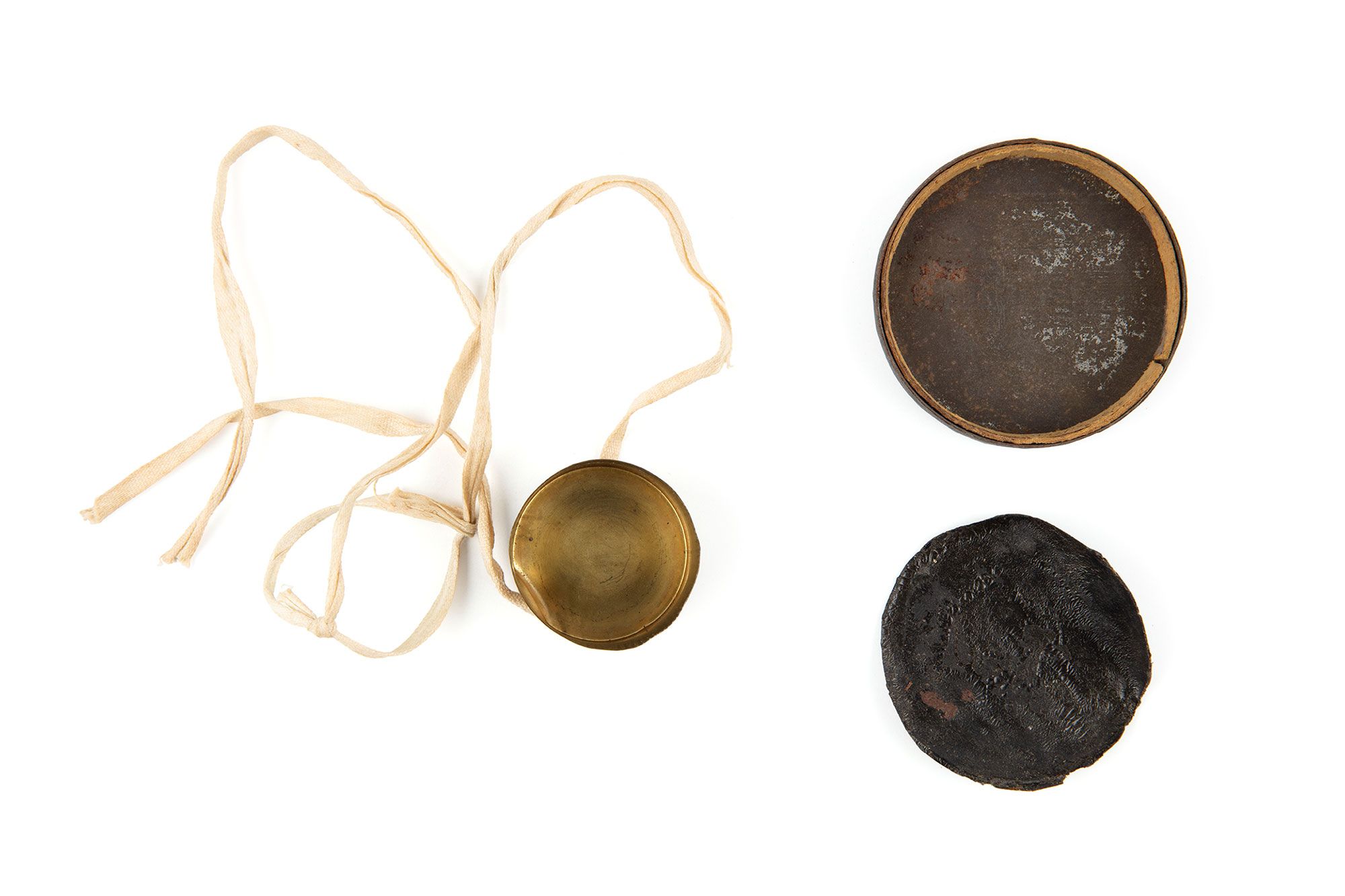 Three round discs of various sizes laid flat. The smallest is brass lens cap with a cream ribbon attached to it. The second largest is black leather.