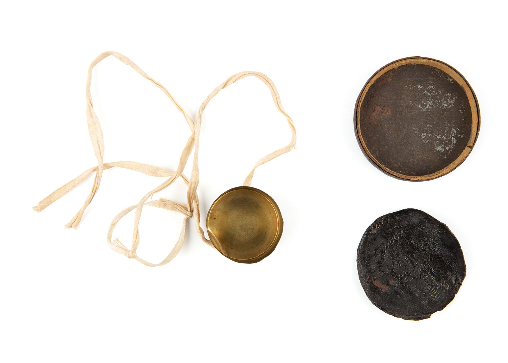 Three round discs of various sizes laid flat. The smallest is brass lens cap with a cream ribbon attached to it. The second largest is black leather.
