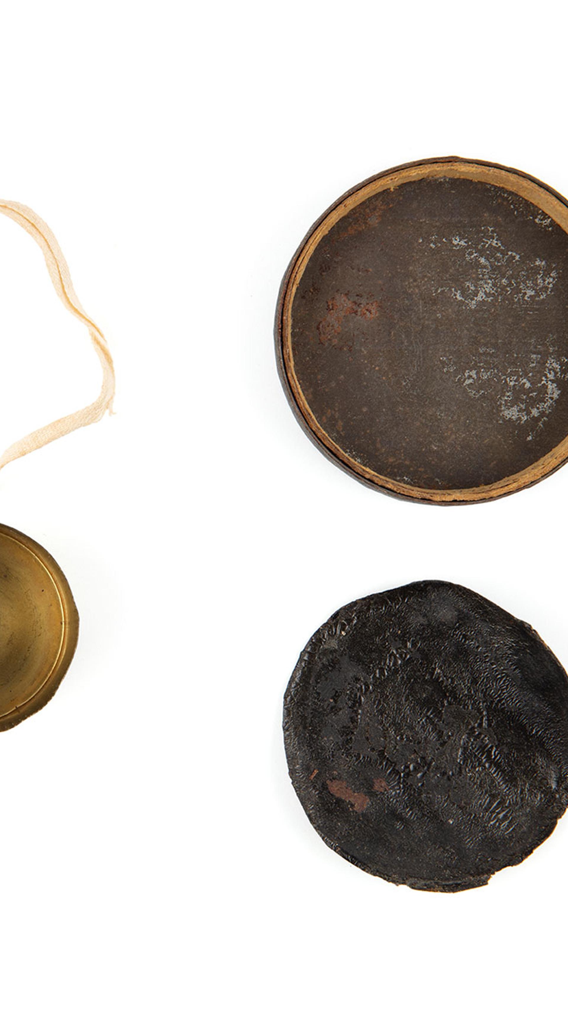 Three round discs of various sizes laid flat. The smallest is brass lens cap with a cream ribbon attached to it. The second largest is black leather.