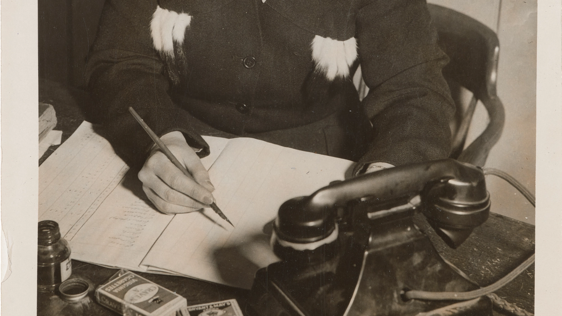 Photograph of a woman sitting at a desk writing, holding a fountain pen, and a Bakelite telephone in front of her.