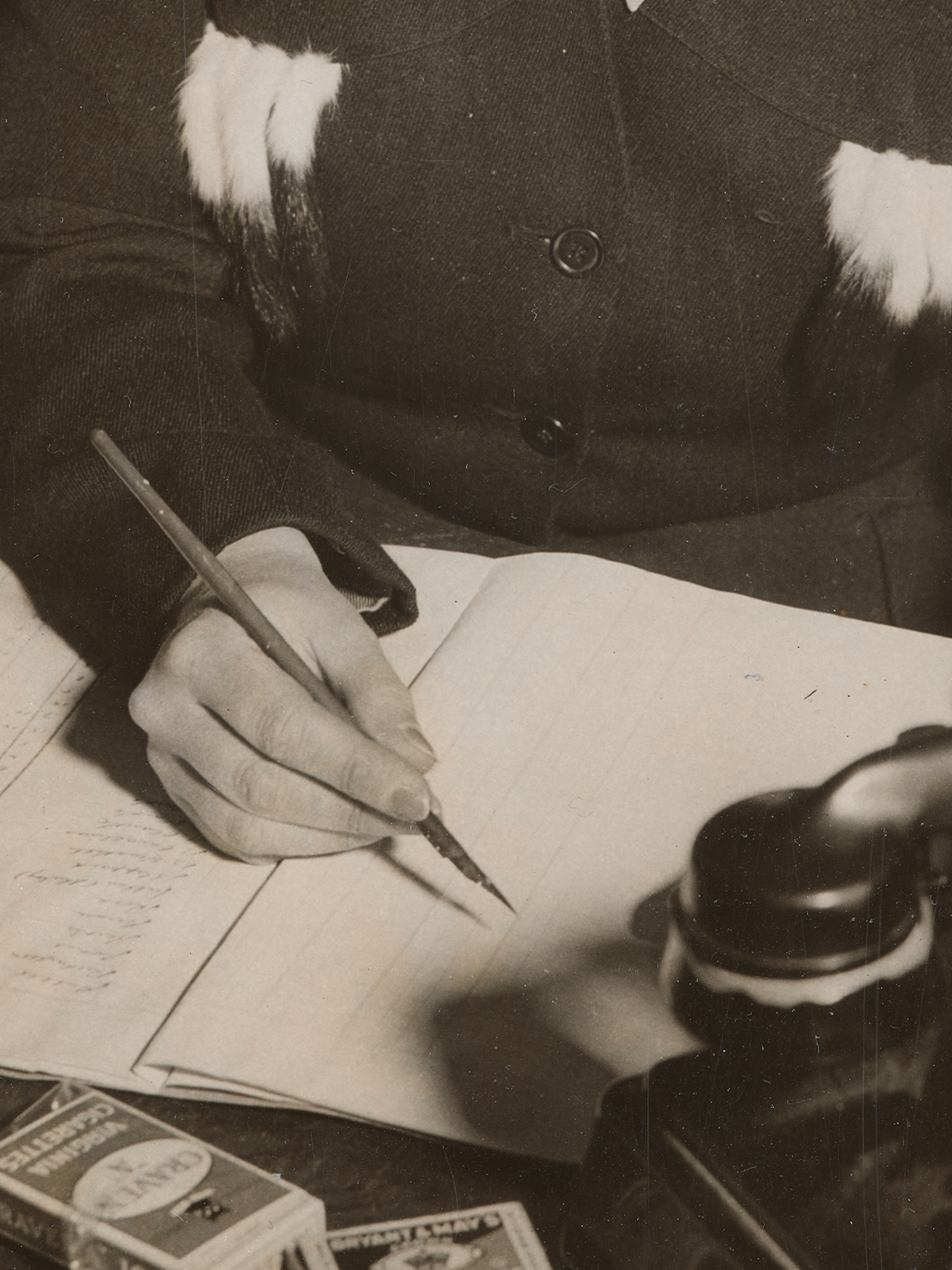 Photograph of a woman sitting at a desk writing, holding a fountain pen, and a Bakelite telephone in front of her.