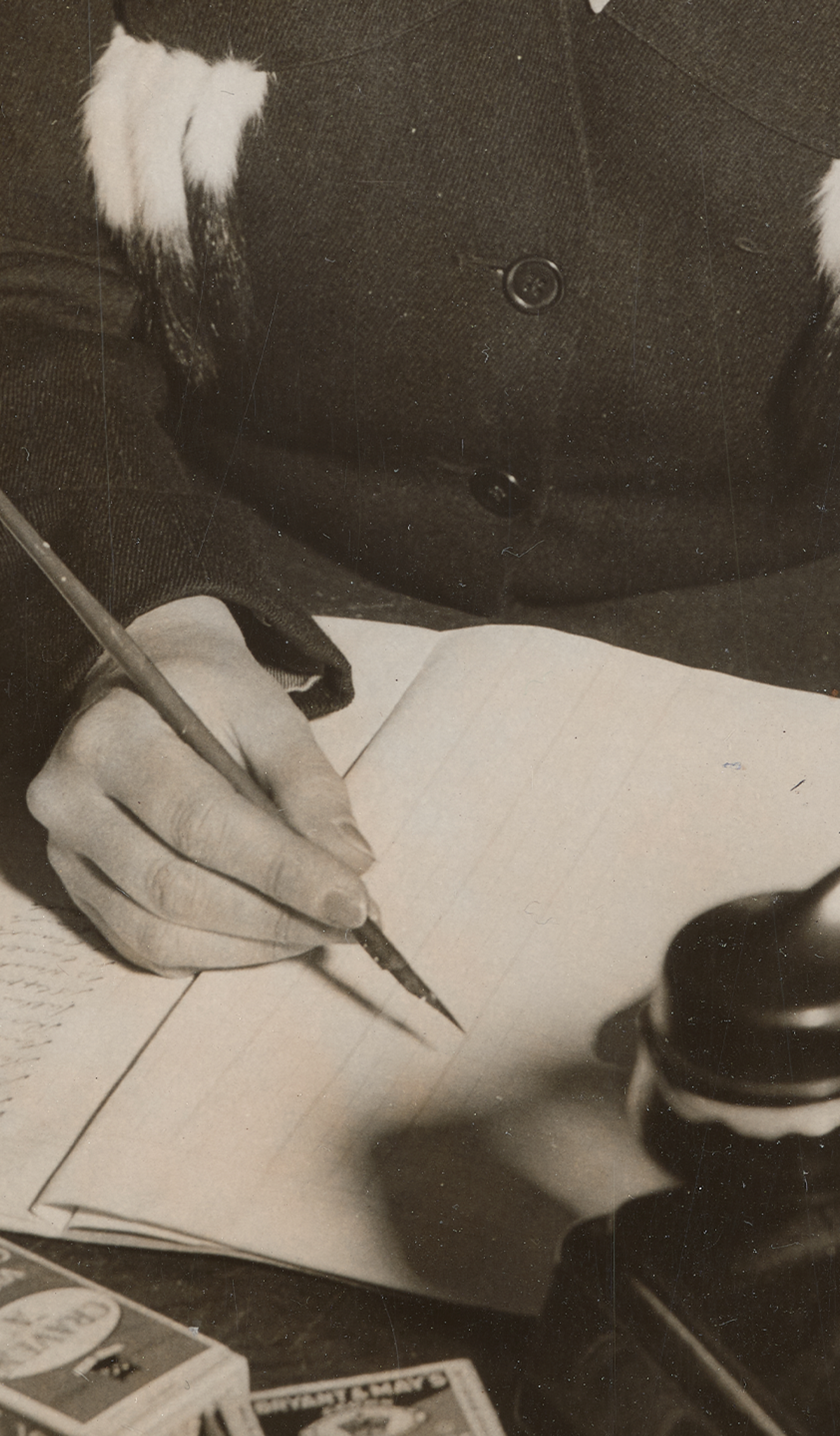 Photograph of a woman sitting at a desk writing, holding a fountain pen, and a Bakelite telephone in front of her.