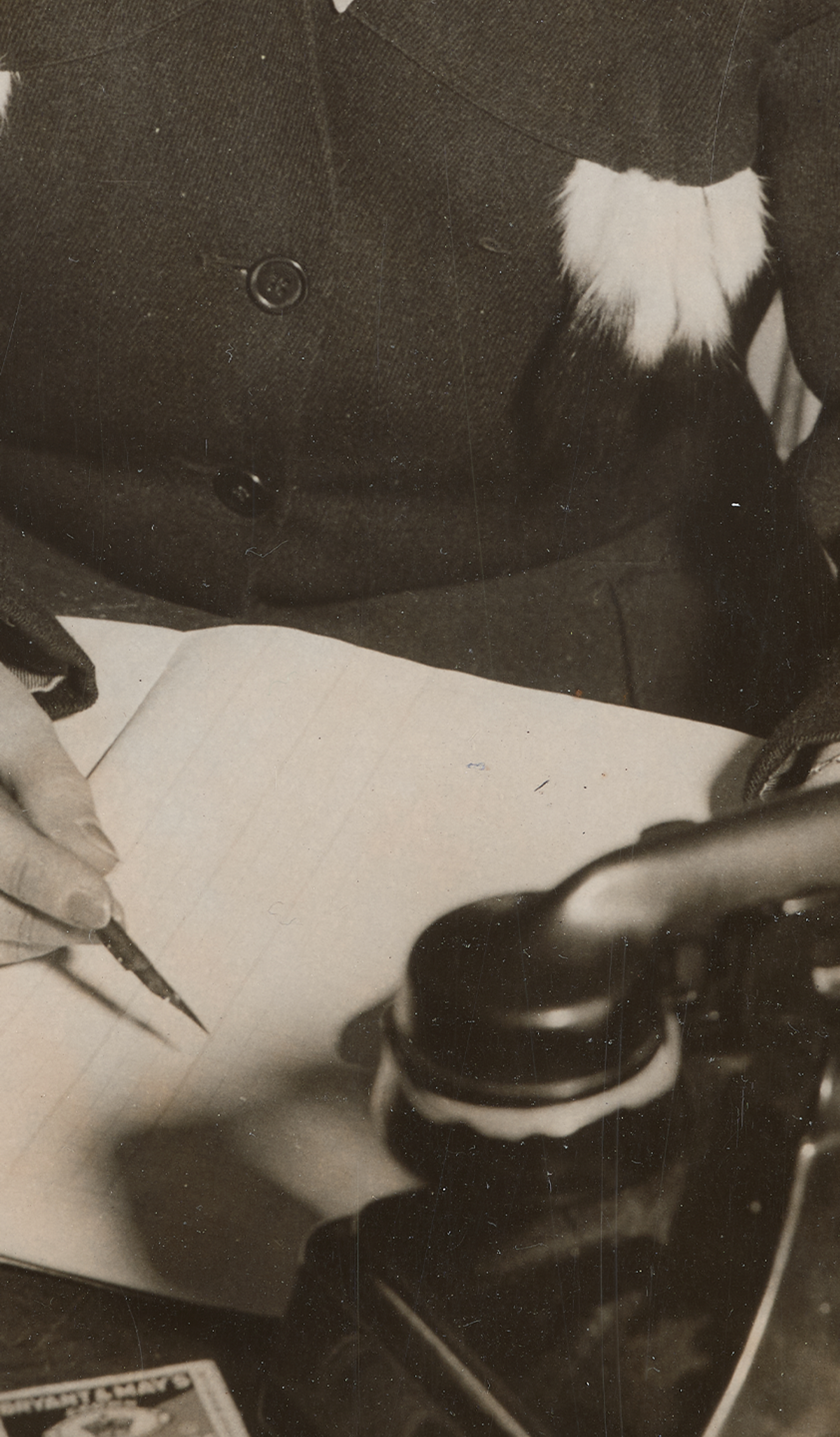 Photograph of a woman sitting at a desk writing, holding a fountain pen, and a Bakelite telephone in front of her.