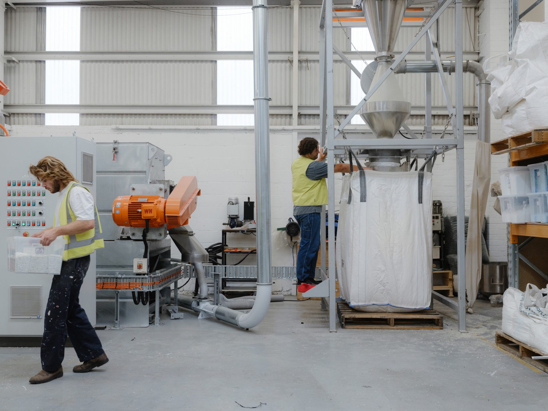 Two figures are working inside a workshop with large pipes and machines surrounding them.