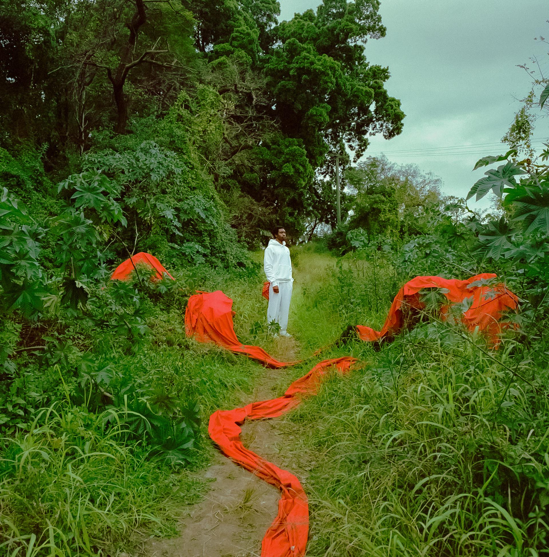 A young man stands in the middle of a forest-like area. He is wearing a white tracksuit. There is a long piece of red fabric draped along the bushes in front of him.
