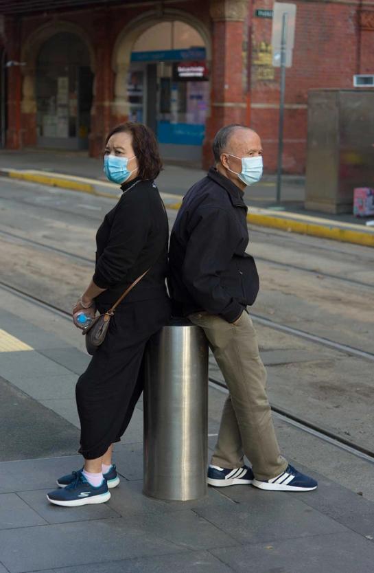 Two people lead back to back on a silver street bollard. They are both wearing navy and white sneakers, black shirts and blue masks.