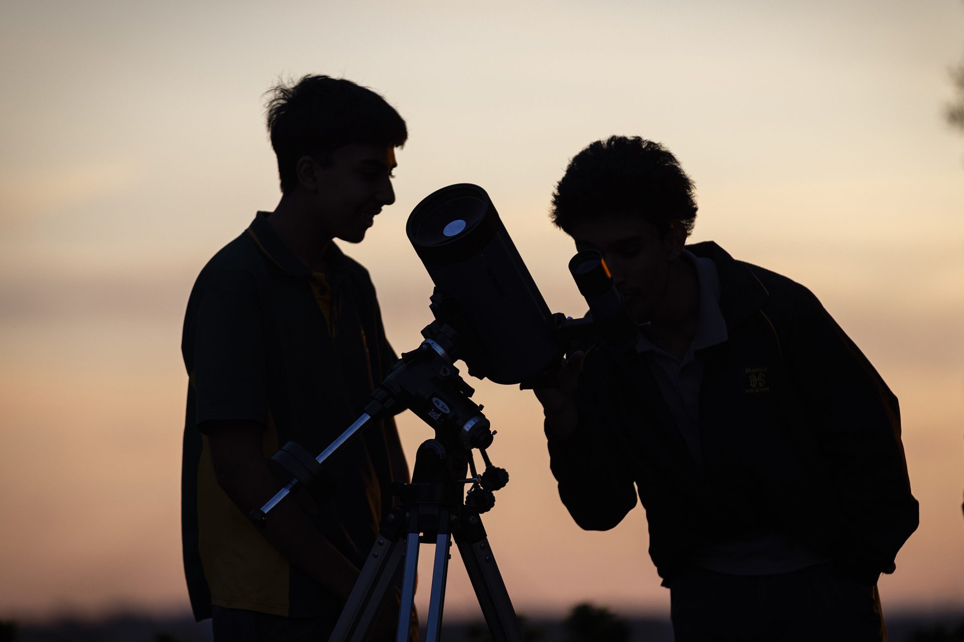 Silhouette of two school boys looking through a telescope, with a pink sunset in the background.