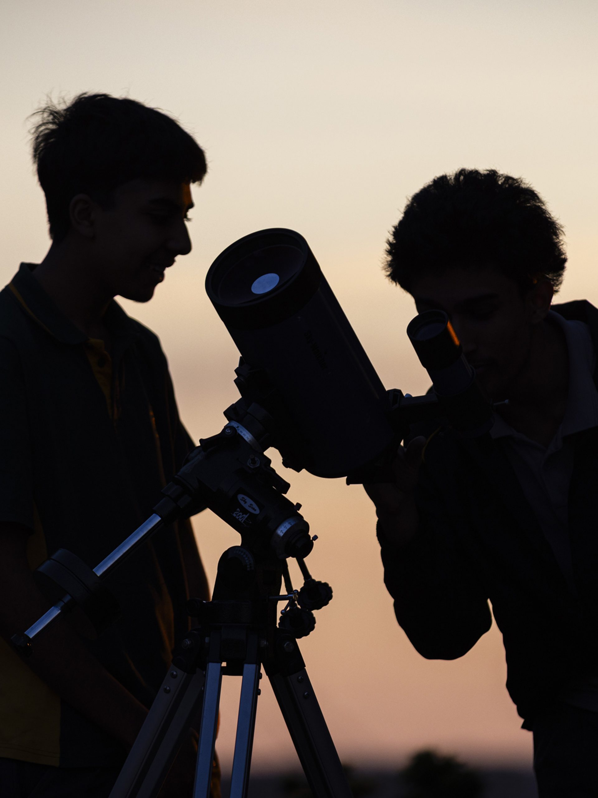 Silhouette of two school boys looking through a telescope, with a pink sunset in the background.
