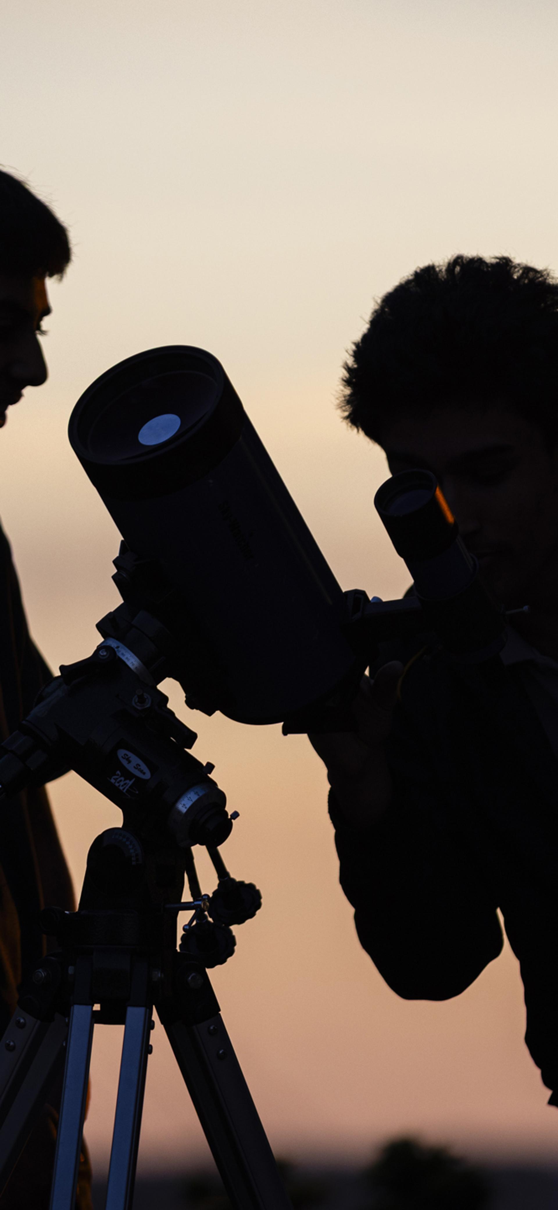Silhouette of two school boys looking through a telescope, with a pink sunset in the background.