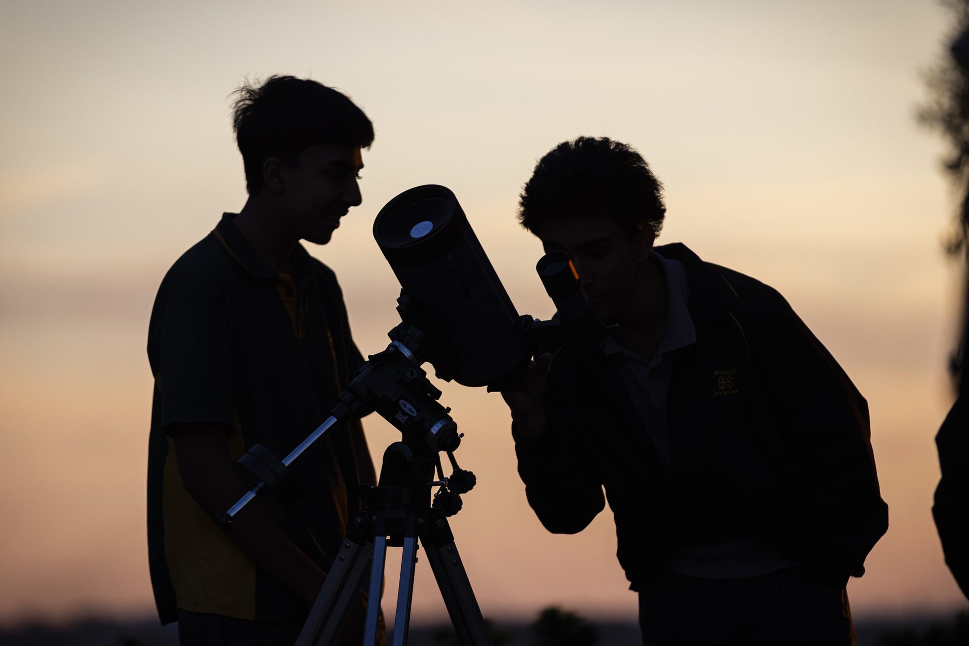 Silhouette of two school boys looking through a telescope, with a pink sunset in the background.