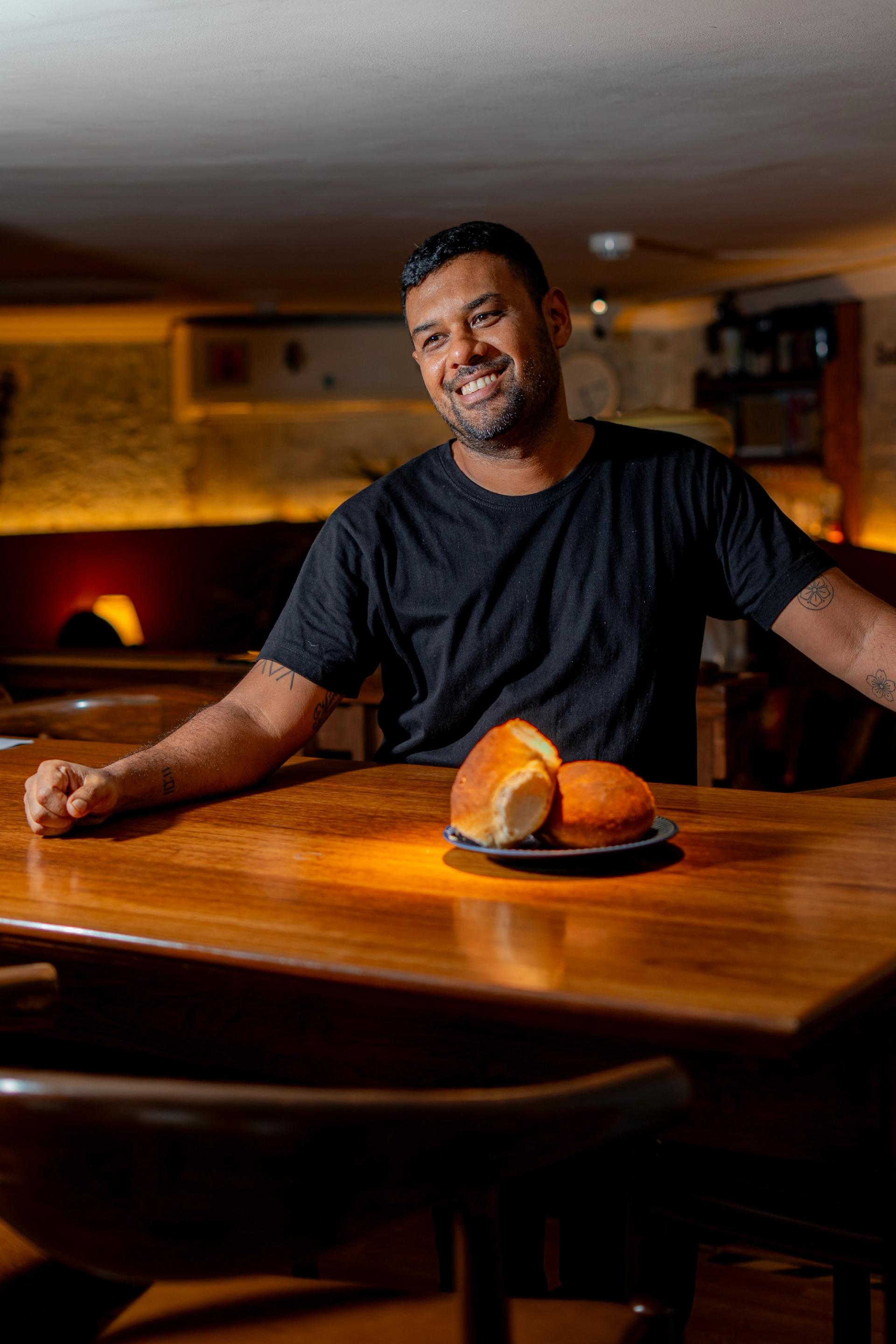 In an empty restaurant, a man stands behind a wooden table. There is a plate of bread rolls in front of him. He wears a black T-shirt and smiles.
