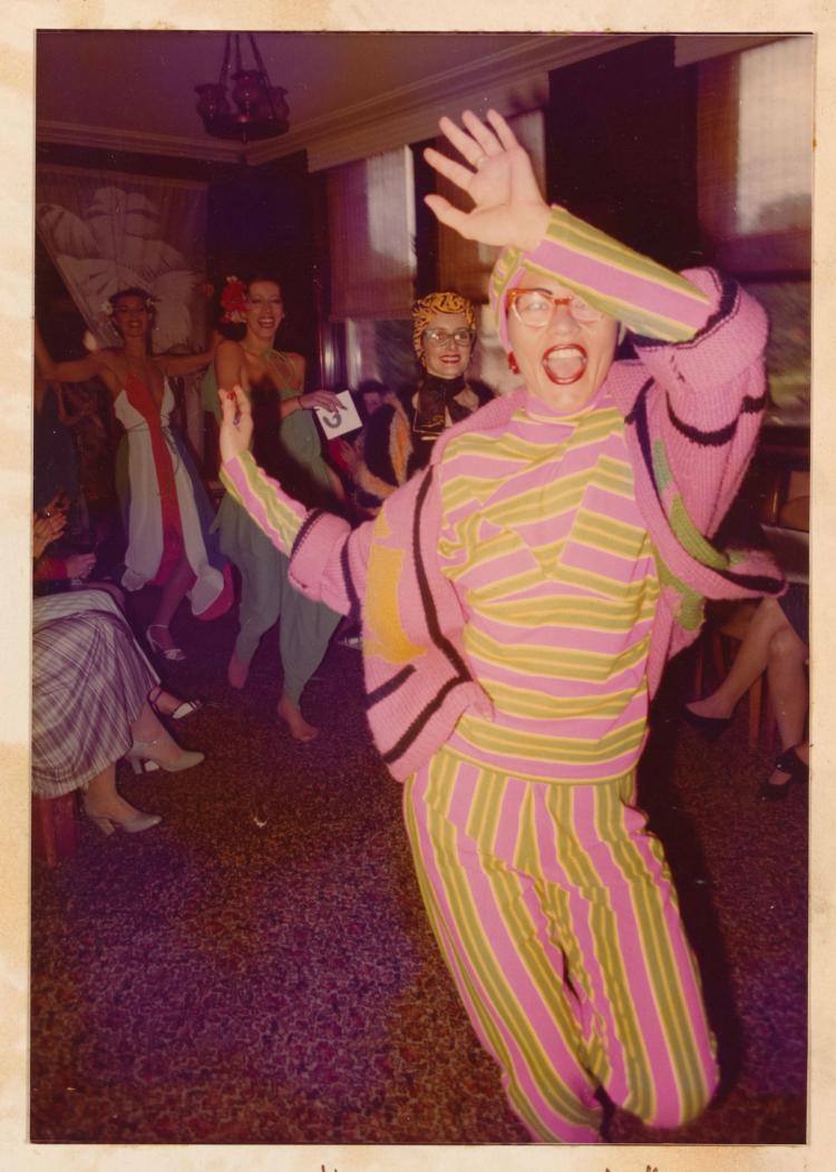 Colour photo of Jenny Kee running towards the camera wearing pink and yellow striped outfit followed by other models in carpeted room