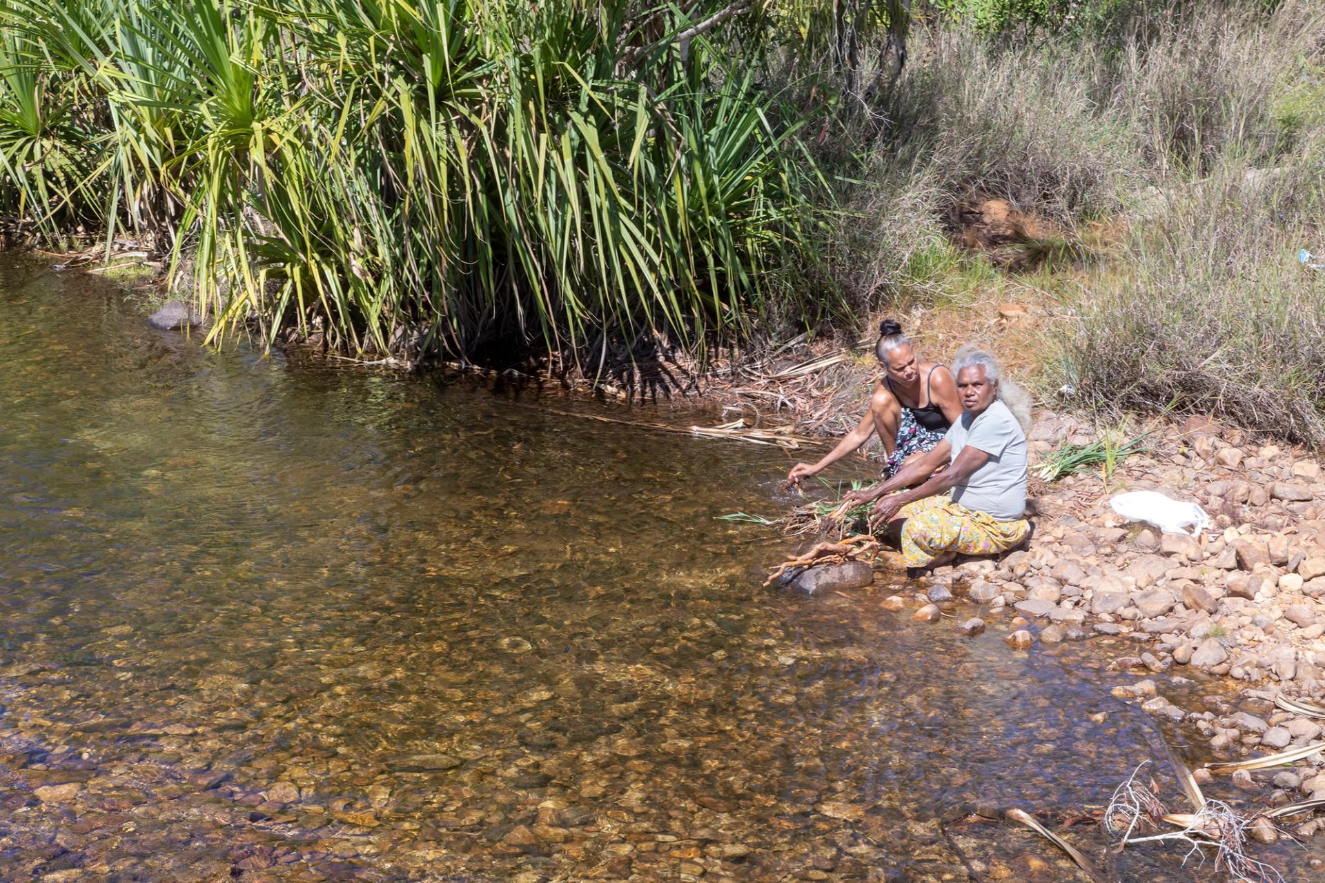 Two women sit on the rocky bed of a river.