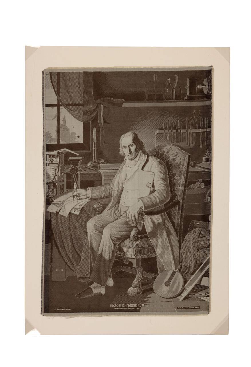 The monochrome portrait of a man sitting in a high-backed patterned arm chair next to a table where he is holding what appears to be a compass over some cards with holes in them - ie punched cards. Behind the chair tools can be seen in a rack on the wall, and on the bench below, a book and other scattered items. Next to the punched cards there is a small scale model of a loom, behind that, on the bench, a vice. There is a window on the left side of the image with a see-through curtain, the vague image of a church spire. Next to the chair on the floor is a pile of fabric and a wheel perhaps from a pulley. The subject is Joseph Marie Jacquard and the process of construction is weaving.