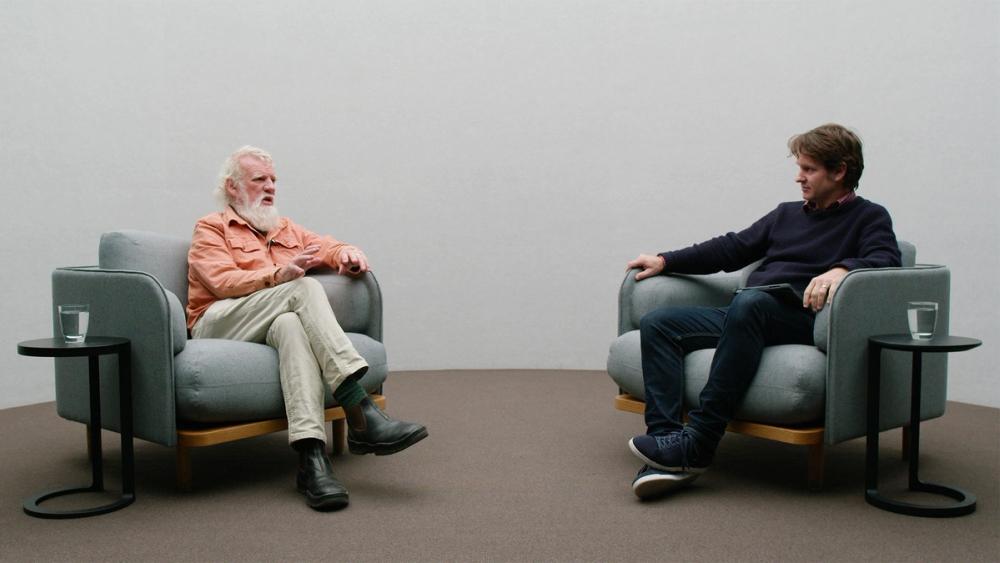 Bruce Pascoe sits in a grey arm chair on the left wearing a peach coloured button up shirt, Craig Reucassel sits on the right in a grey arm chair wearing jeans and a navy jumper.