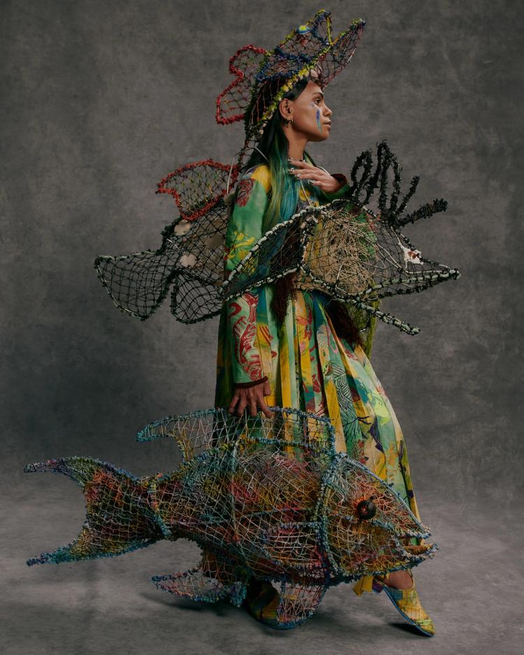 A model poses against a grey background wearing a multicoloured floor-length dress and holding two large coloured woven fish.