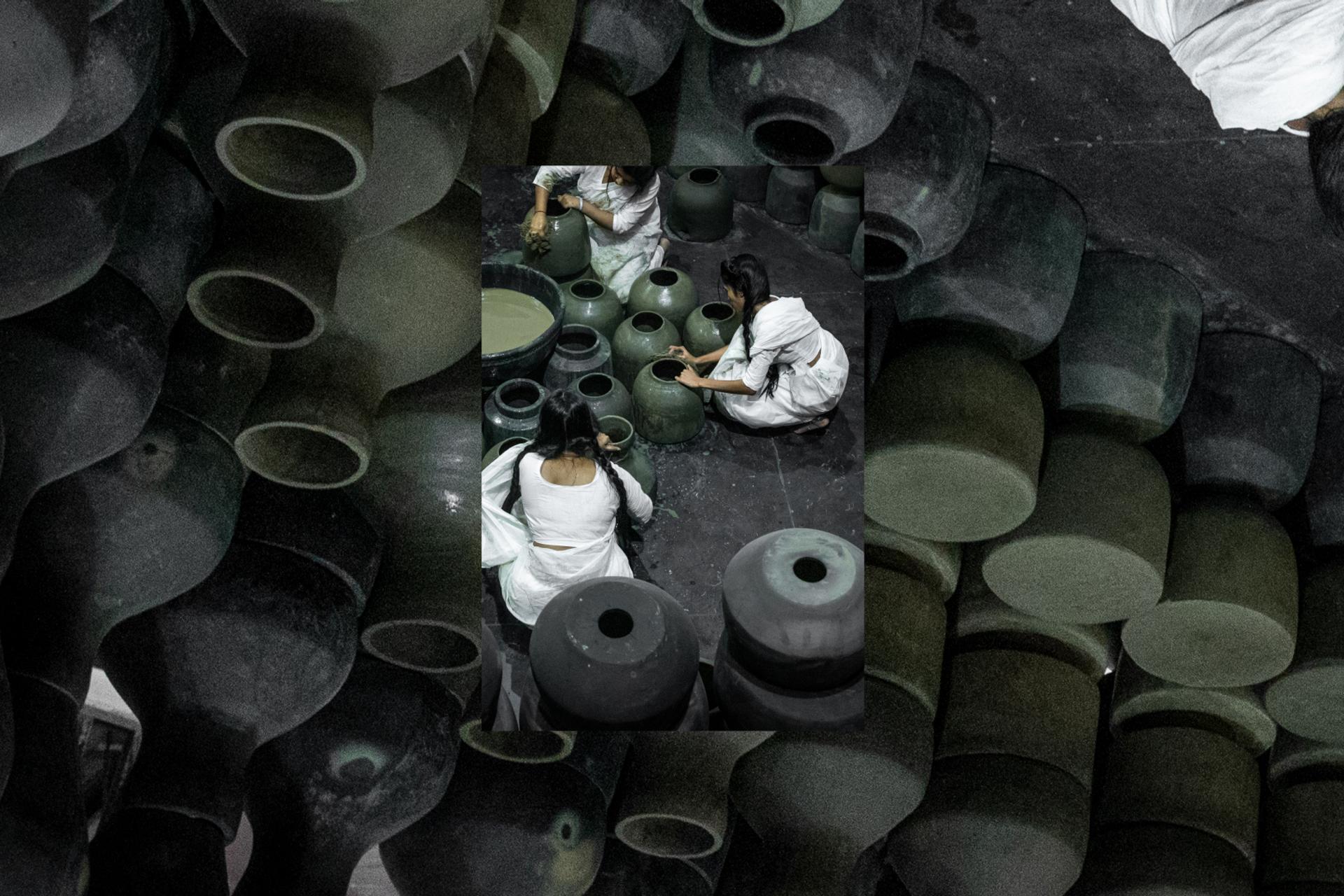 Five people dressed in white sit on the floor around black pots and surrounded by black pots.