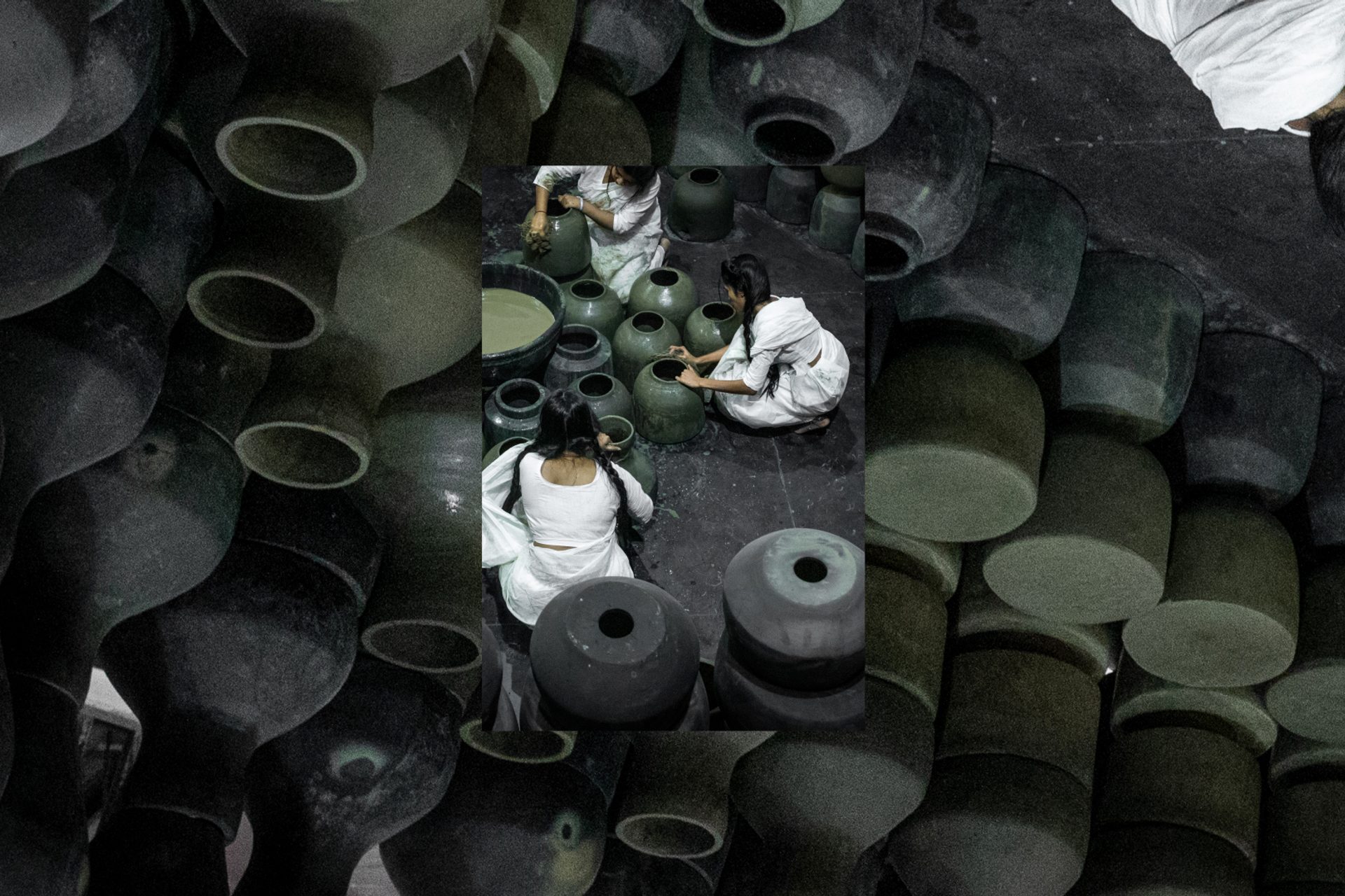 Five people dressed in white sit on the floor around black pots and surrounded by black pots.