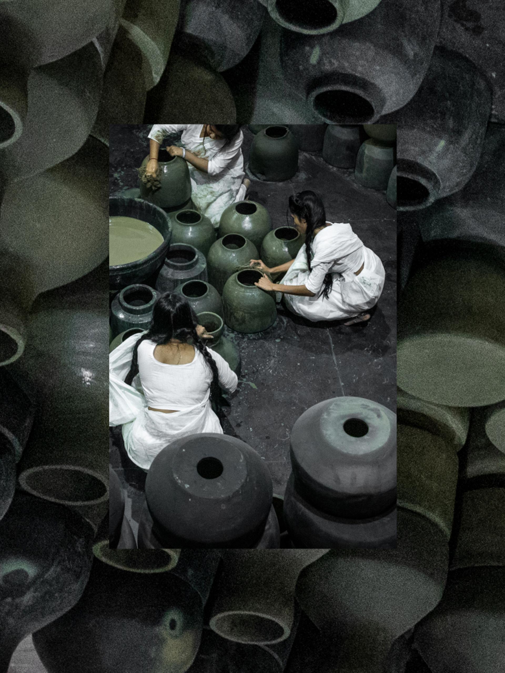 Five people dressed in white sit on the floor around black pots and surrounded by black pots.