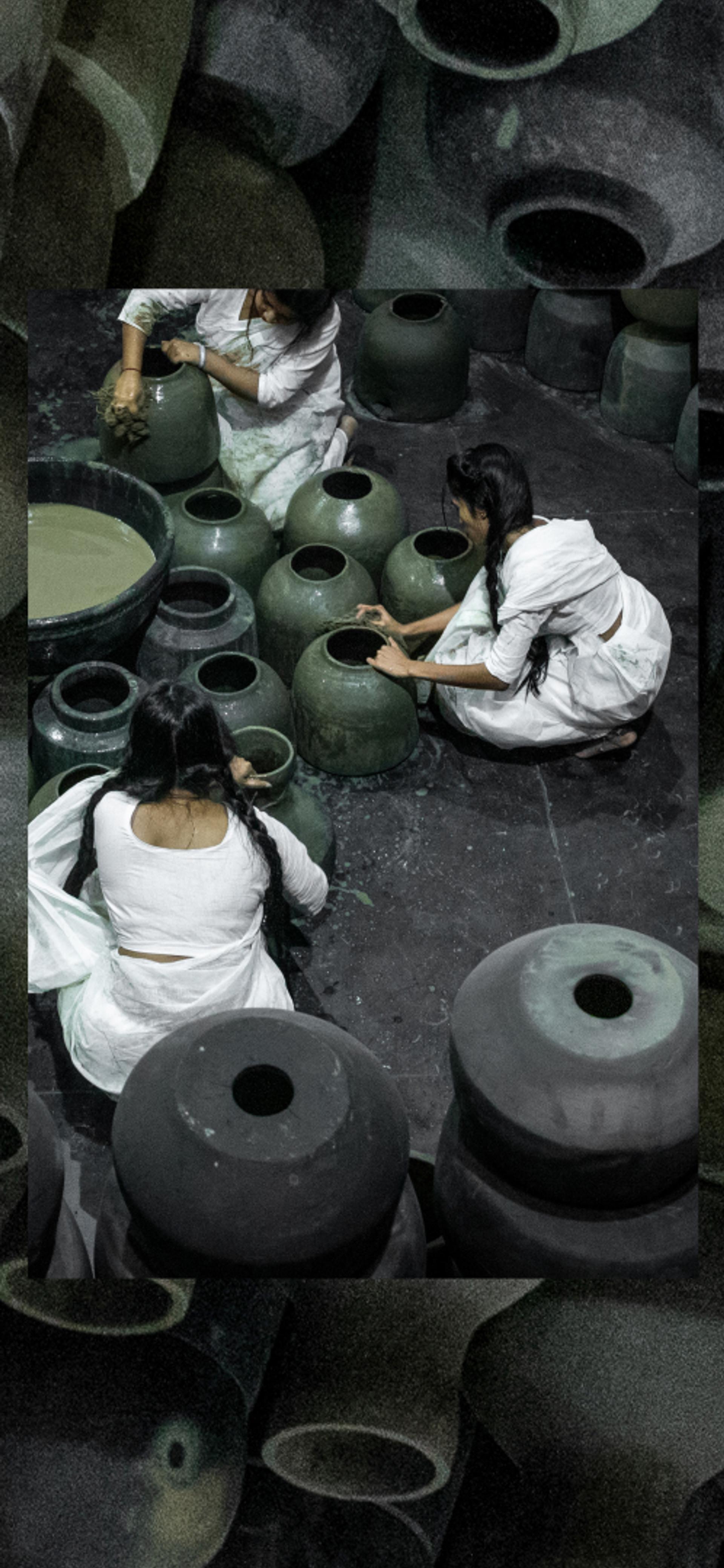 Five people dressed in white sit on the floor around black pots and surrounded by black pots.