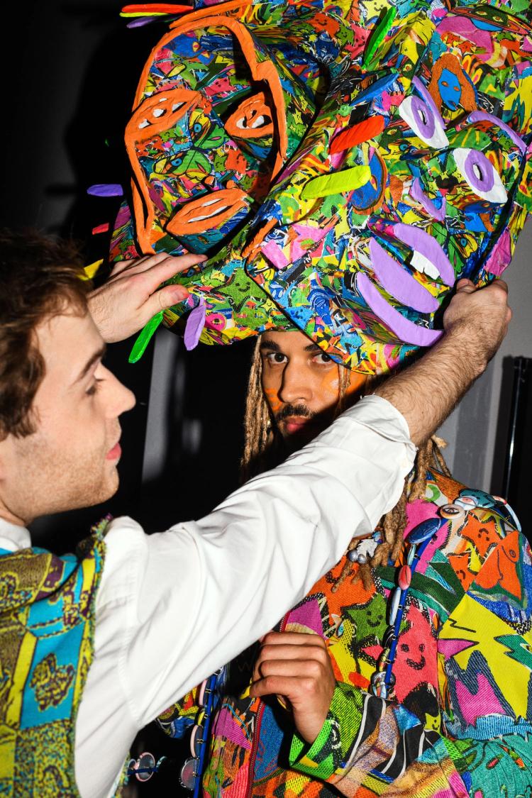 Max Rixon puts a multicoloured hat featuring cut out faces on the head of model Ryva