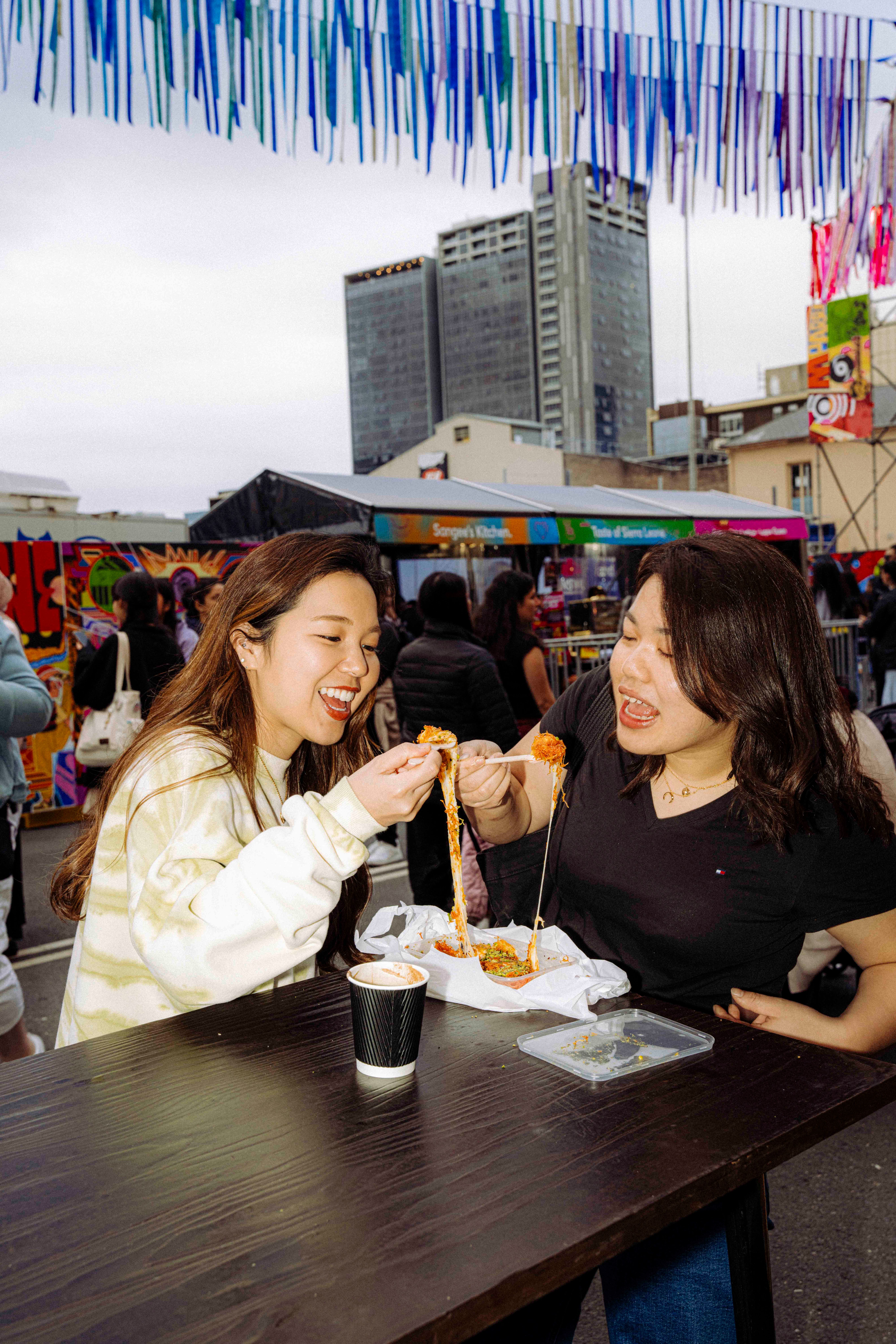 Two smiling young people share a dish at an outdoor market, pulling apart strands of food while seated at a table, with colourful streamer decorations overhead.