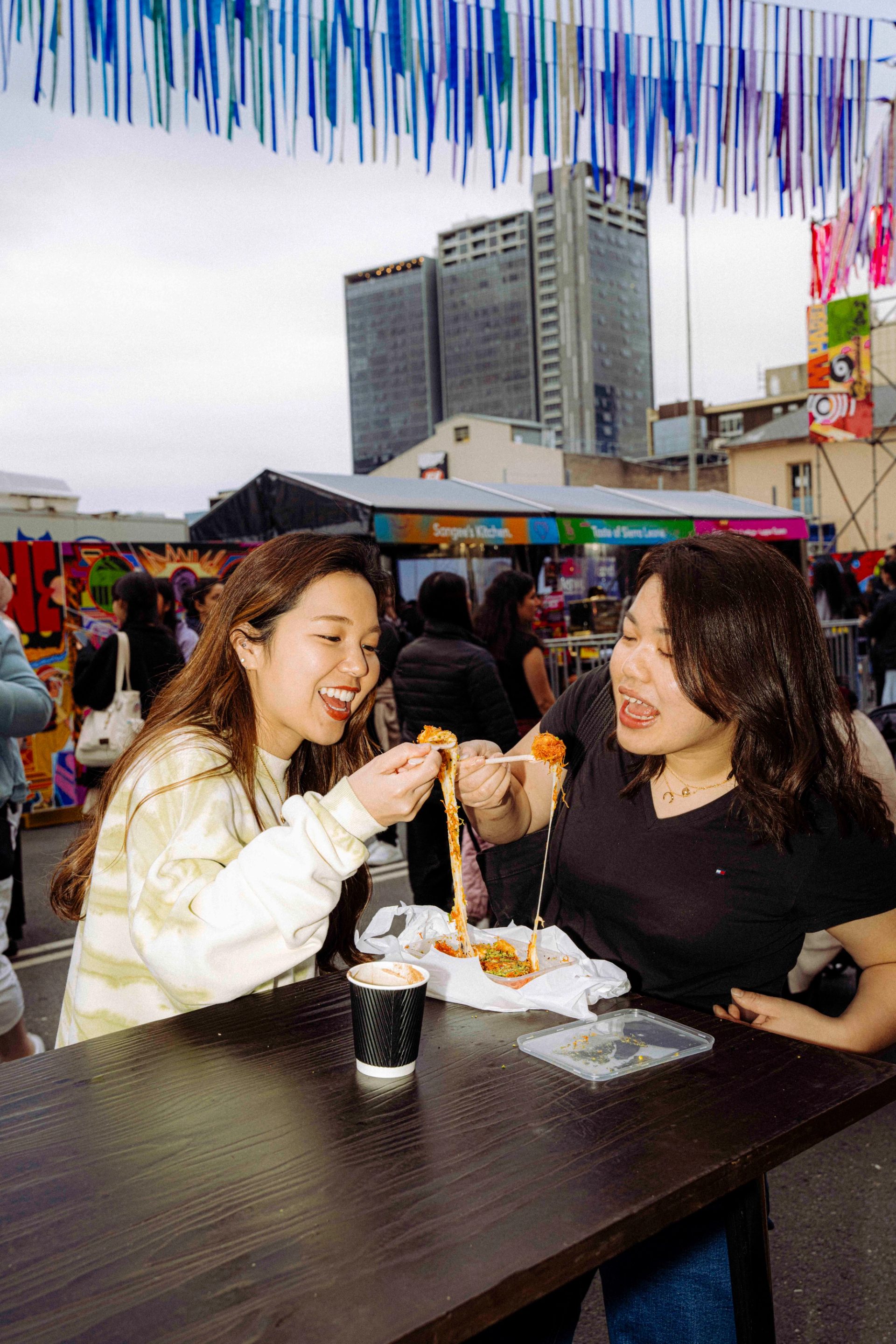 Two smiling young people share a dish at an outdoor market, pulling apart strands of food while seated at a table, with colourful streamer decorations overhead.