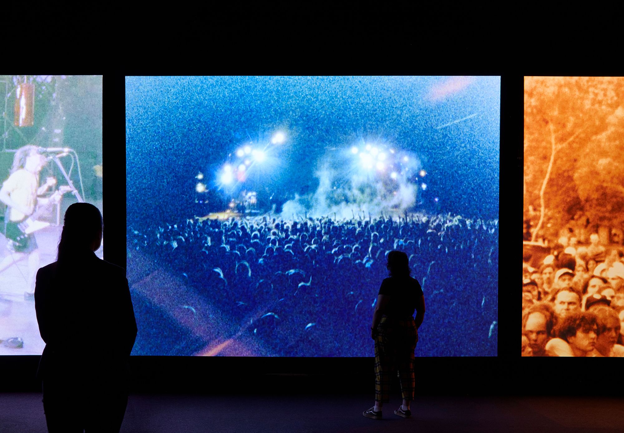Two people watch large screens of music concerts.