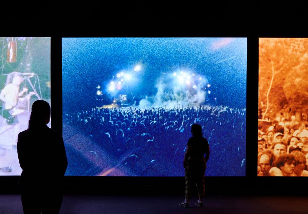 Two people watch large screens of music concerts.