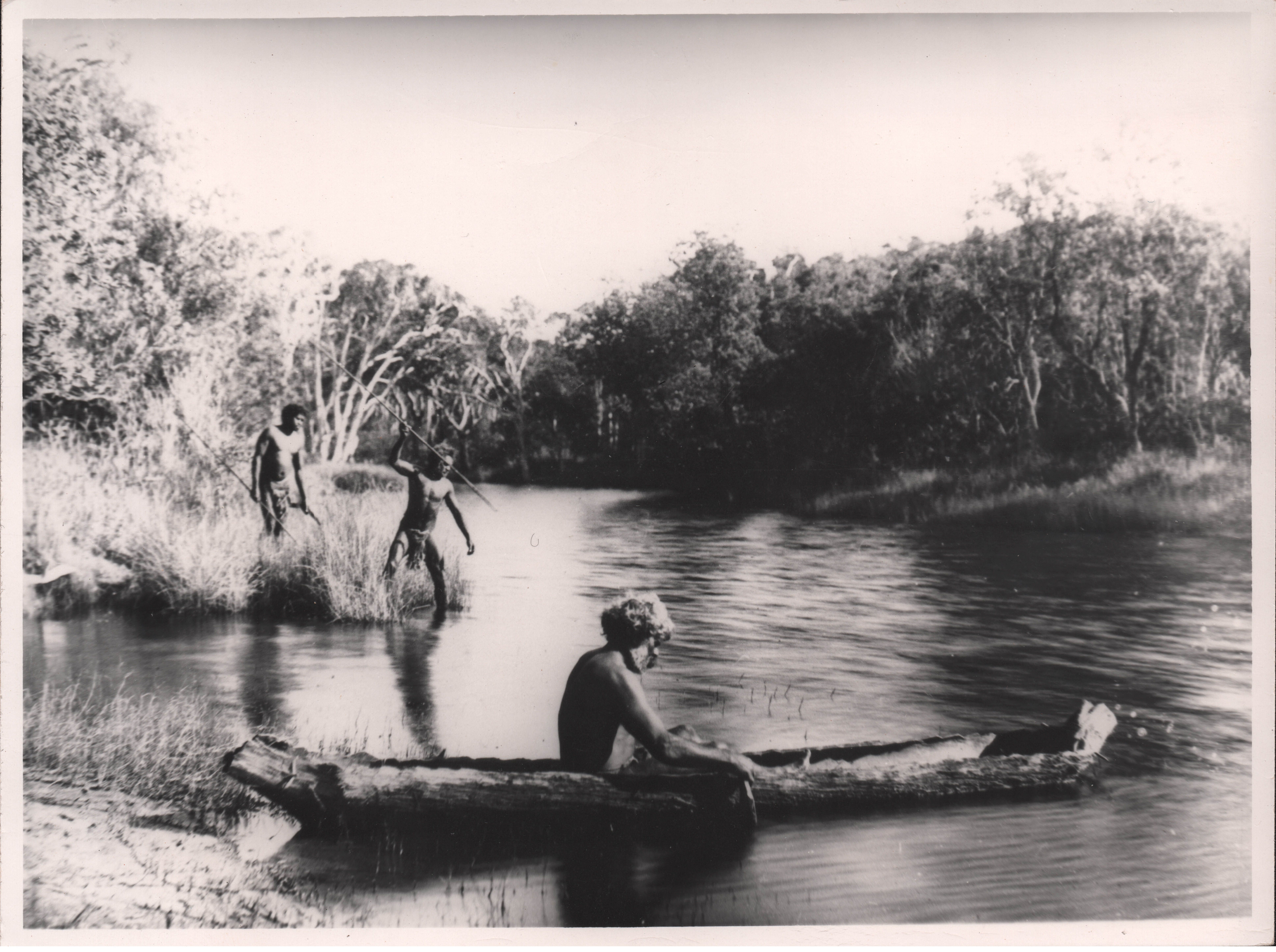 Three Birpai men at a river. One man is in a canoe, one man is spearing fish and another stands behind him.