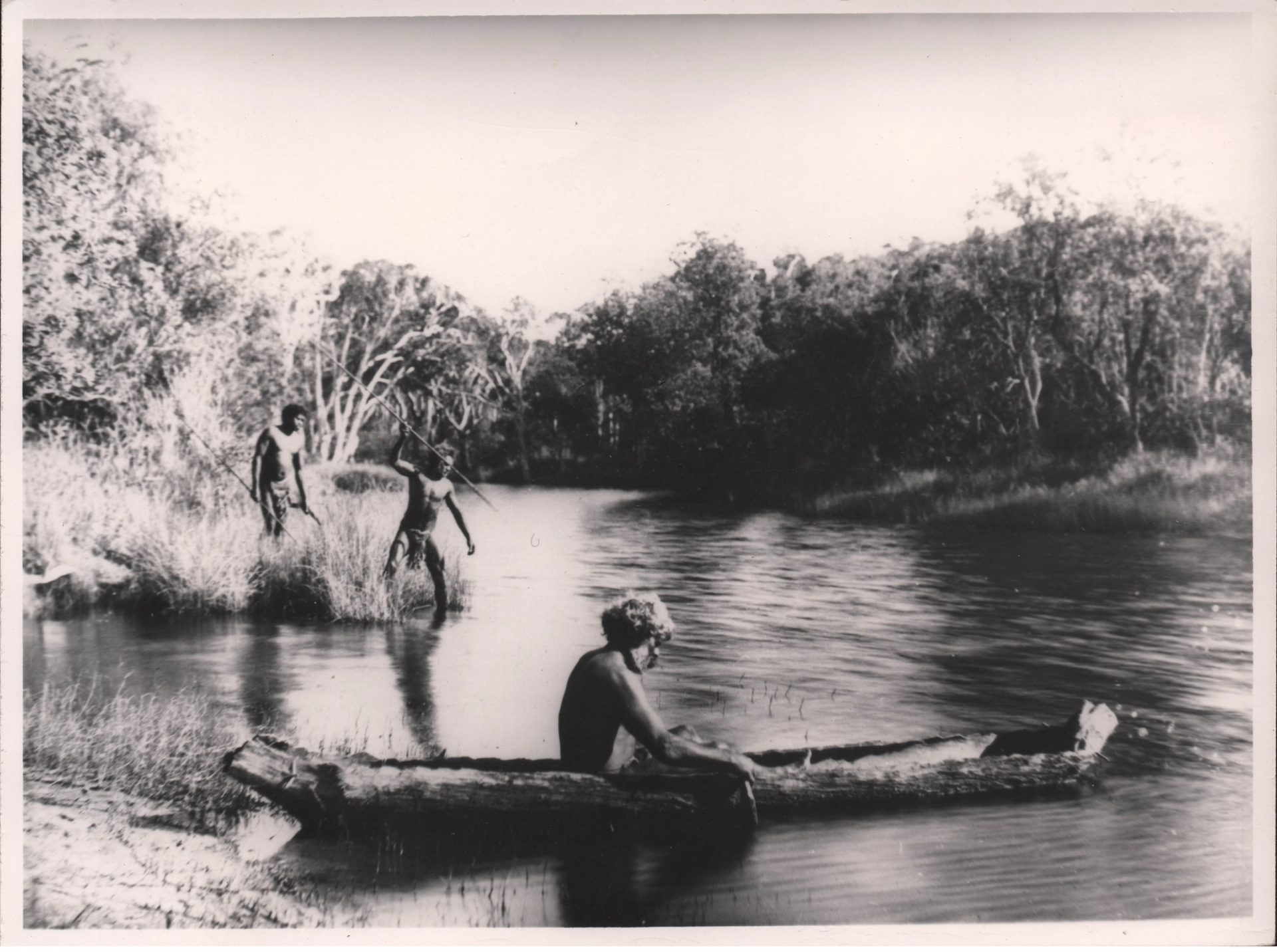 Three Birpai men at a river. One man is in a canoe, one man is spearing fish and another stands behind him.