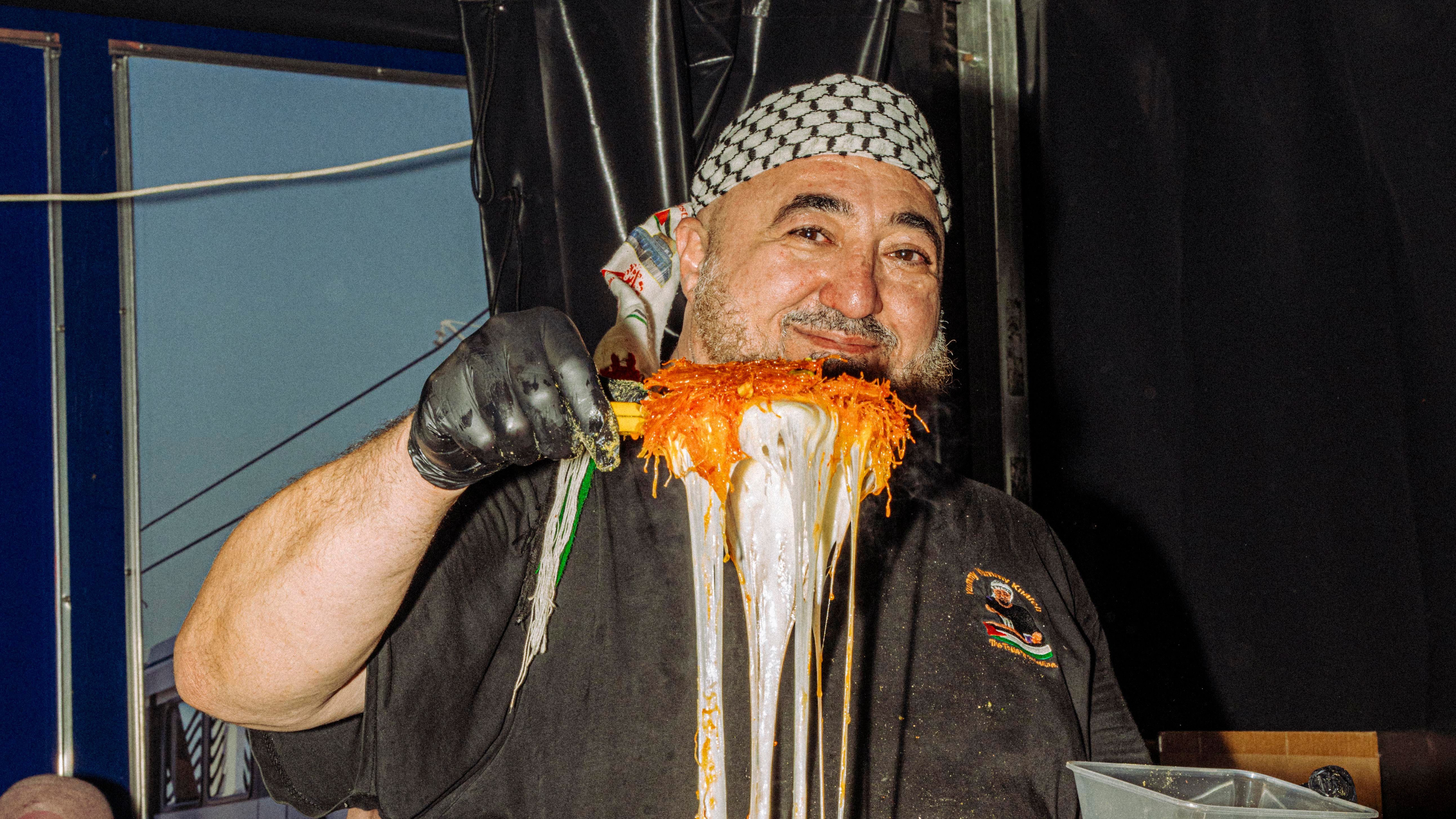 A smiling vendor who is wearing a black shirt and patterned headscarf holds a large metallic tray above their head in a festive food stall setting.
