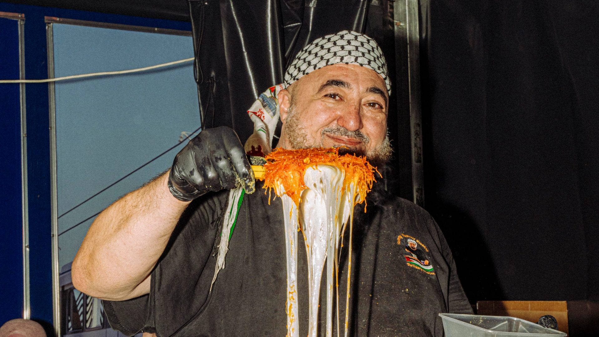 A smiling vendor who is wearing a black shirt and patterned headscarf holds a large metallic tray above their head in a festive food stall setting.