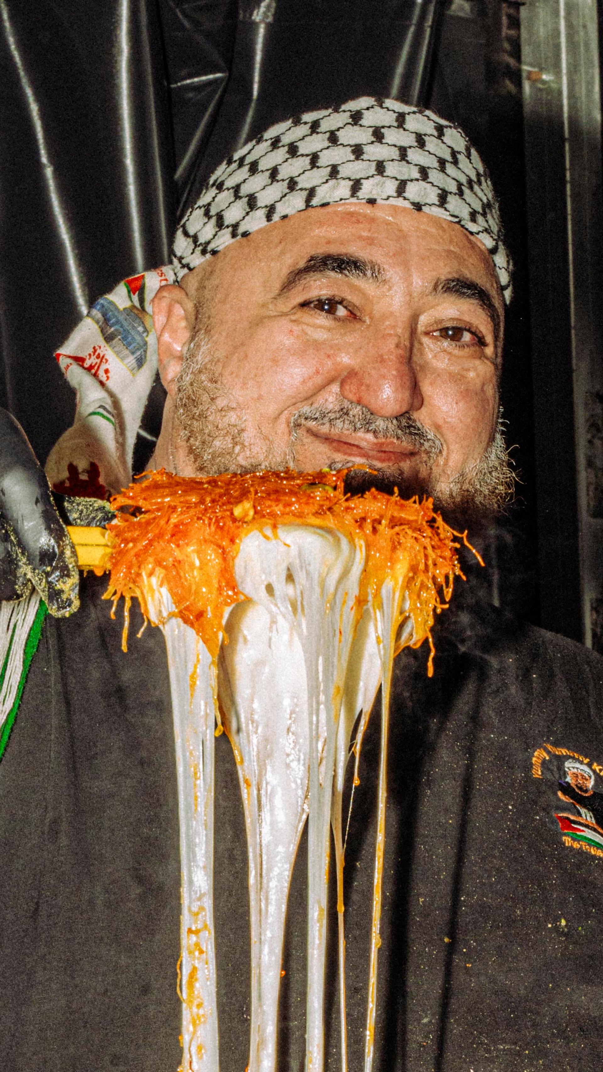 A smiling vendor who is wearing a black shirt and patterned headscarf holds a large metallic tray above their head in a festive food stall setting.