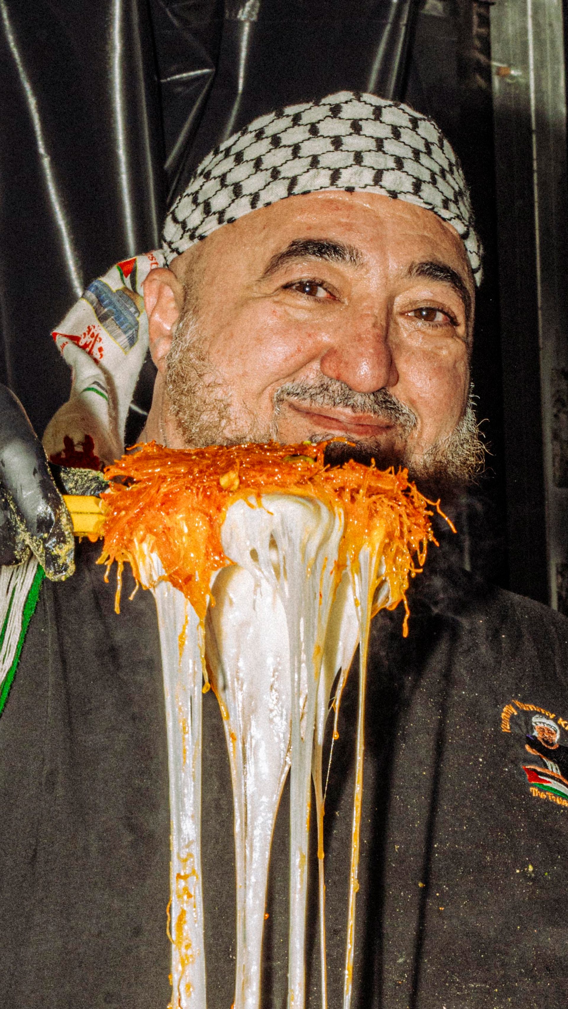 A smiling vendor who is wearing a black shirt and patterned headscarf holds a large metallic tray above their head in a festive food stall setting.