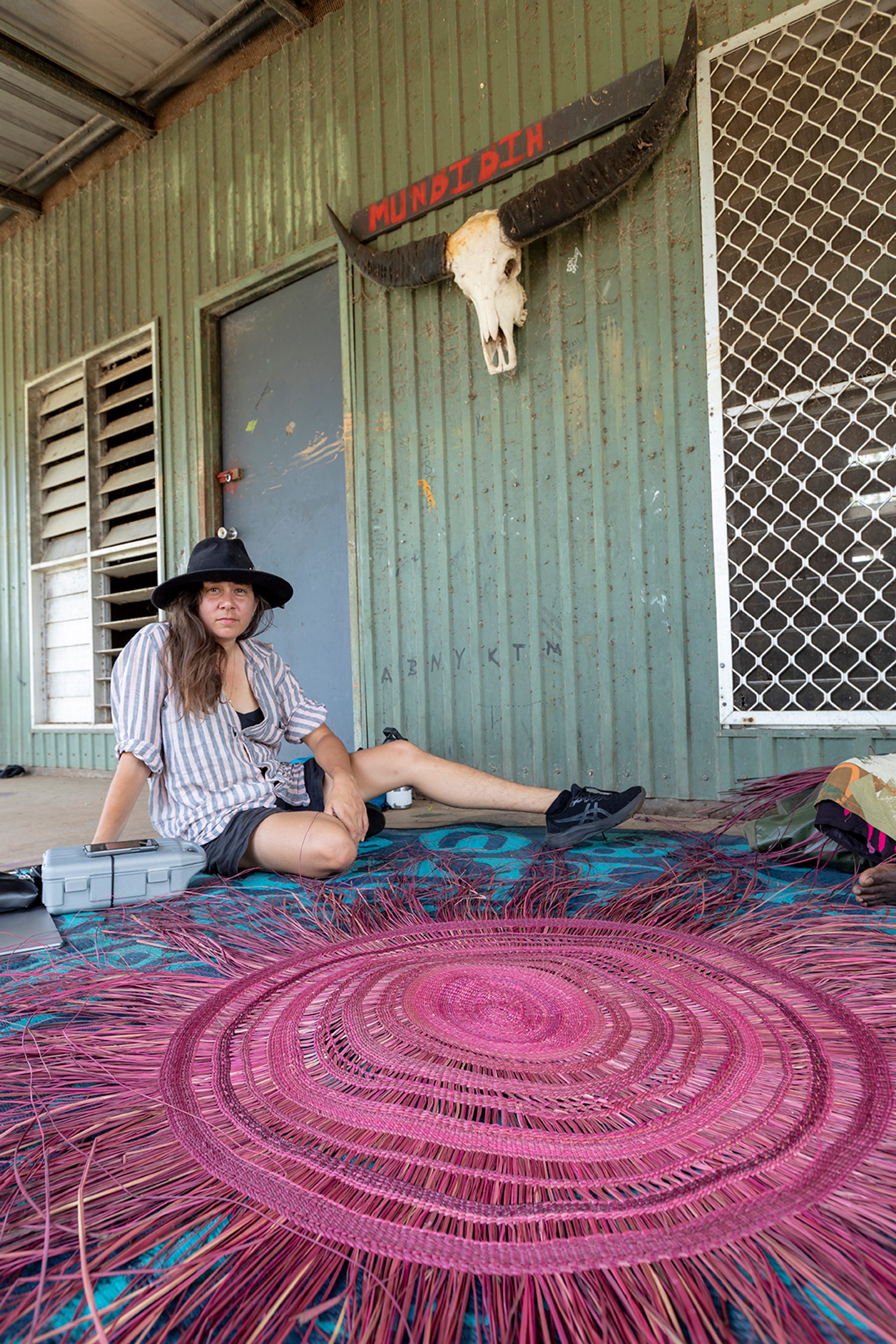 A woman sits outside a shed with faded green corrugated metal walls.