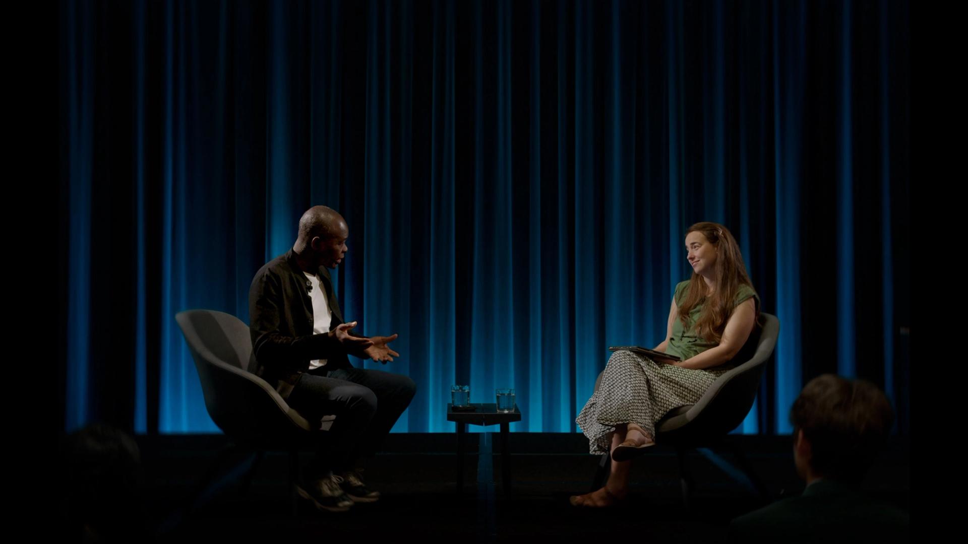 Two figures sitting on chairs in conversation facing each other, a blue draped curtain behind them.