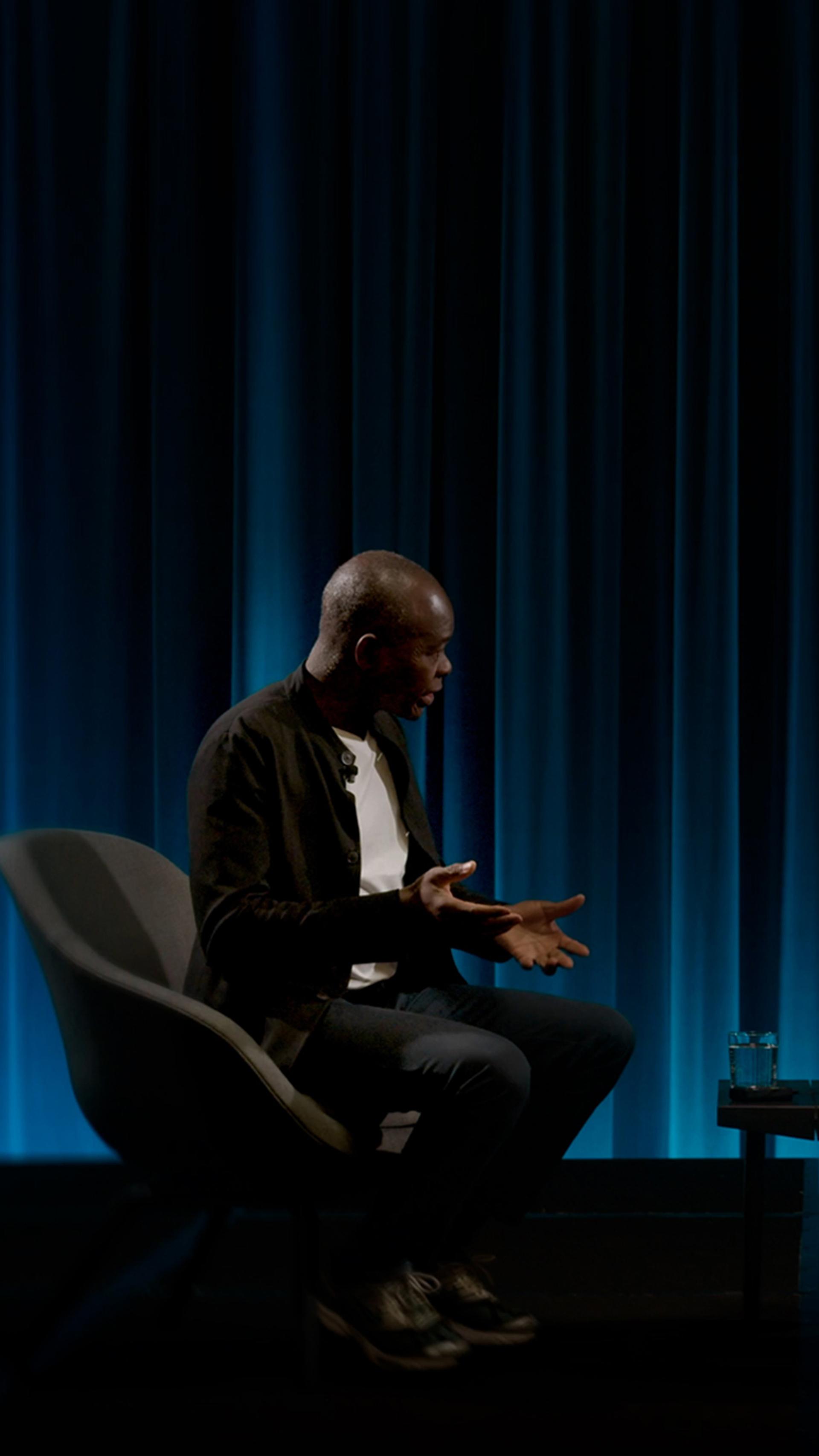 Two figures sitting on chairs in conversation facing each other, a blue draped curtain behind them.