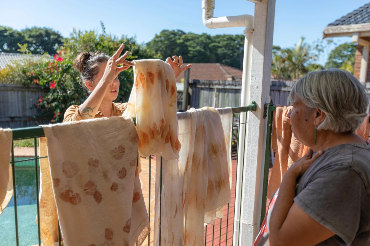 Two women stand on either side of a household pool fence that has several hand dyed textiles draped over it.
